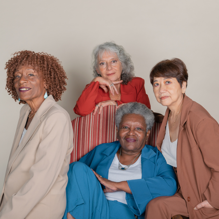 Four mature women posing together against a plain beige background, dressed in colorful professional attire.