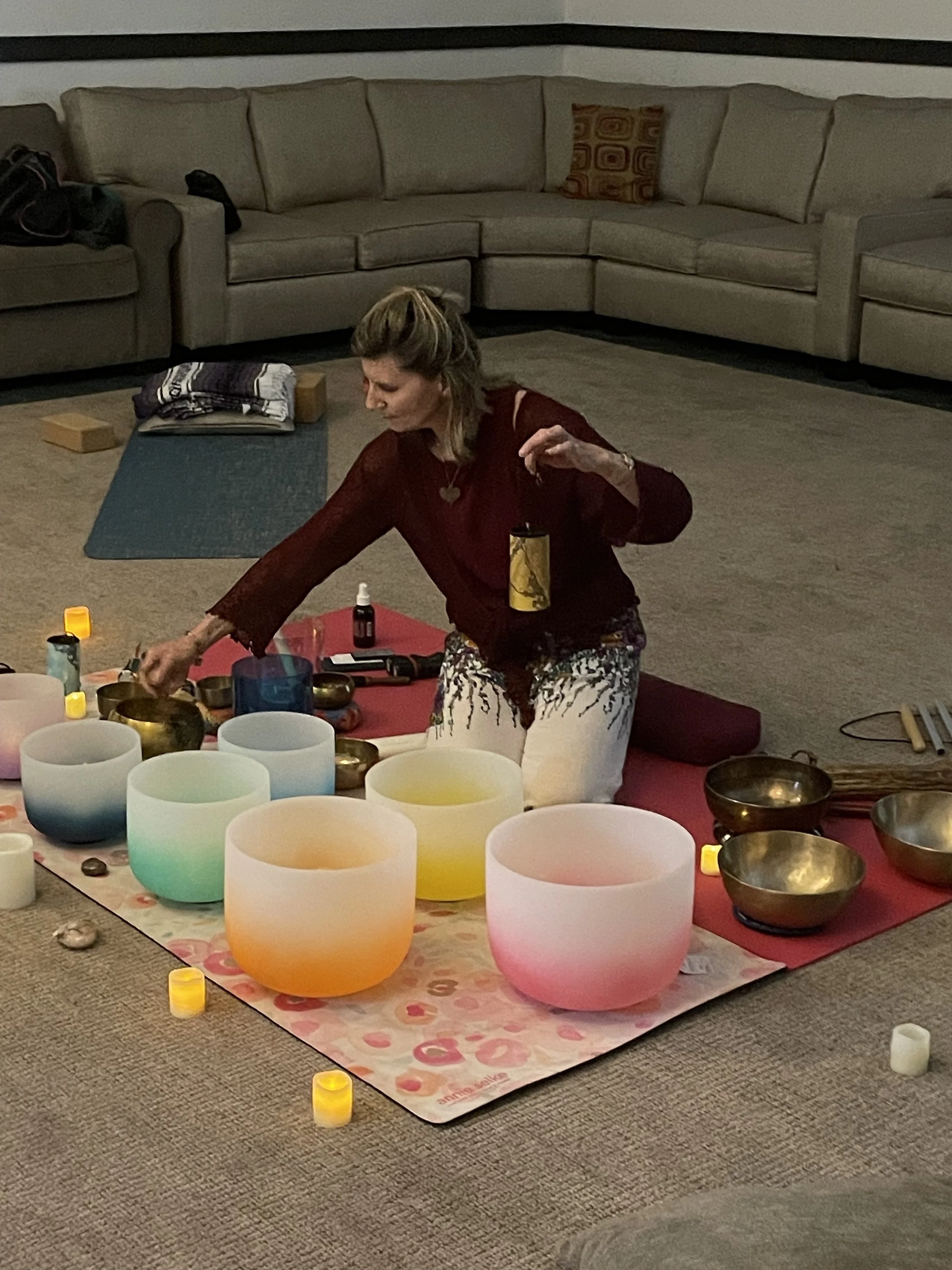 A woman is playing crystal singing bowls on a pink mat in a living room with beige seating, candles, and other meditation items.