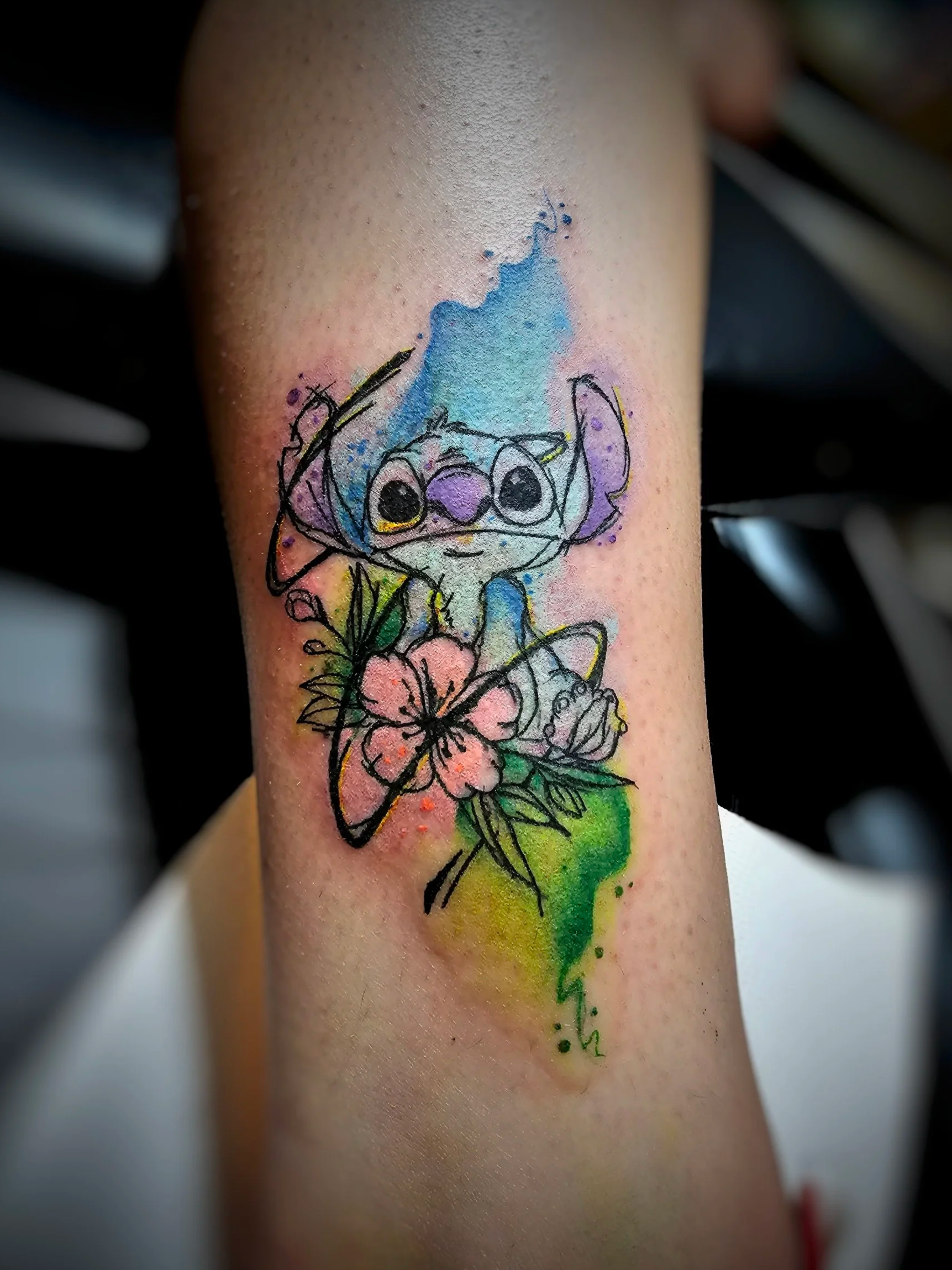 A tattoo on skin of a colorful illustration of Stitch from Disney's Lilo and Stitch, with a blue splash background and a pink hibiscus flower with green leaves below him.