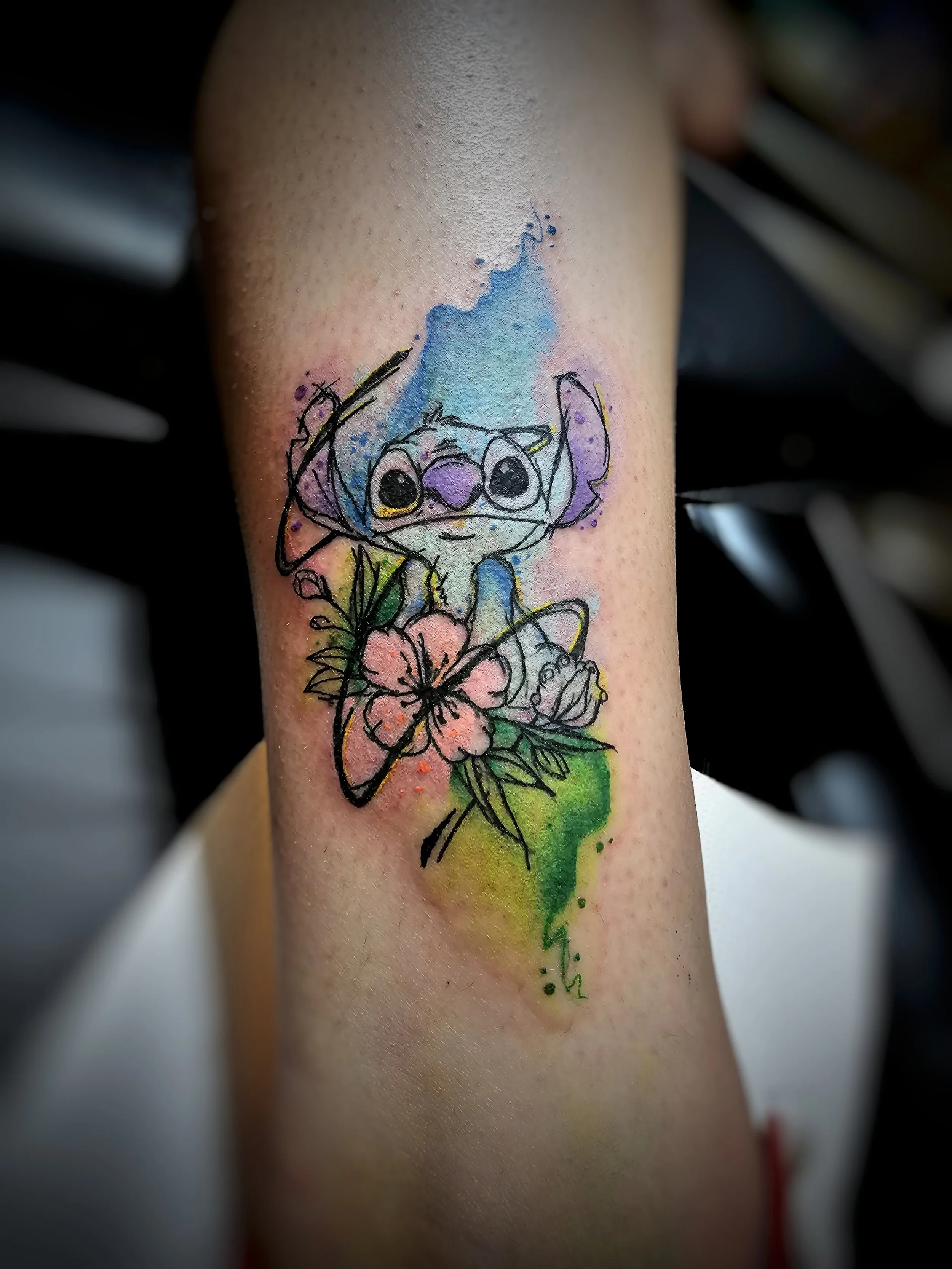 Colorful tattoo of a cartoonish creature with large eyes, surrounded by flowers, with watercolor splashes and a rainbow background.
