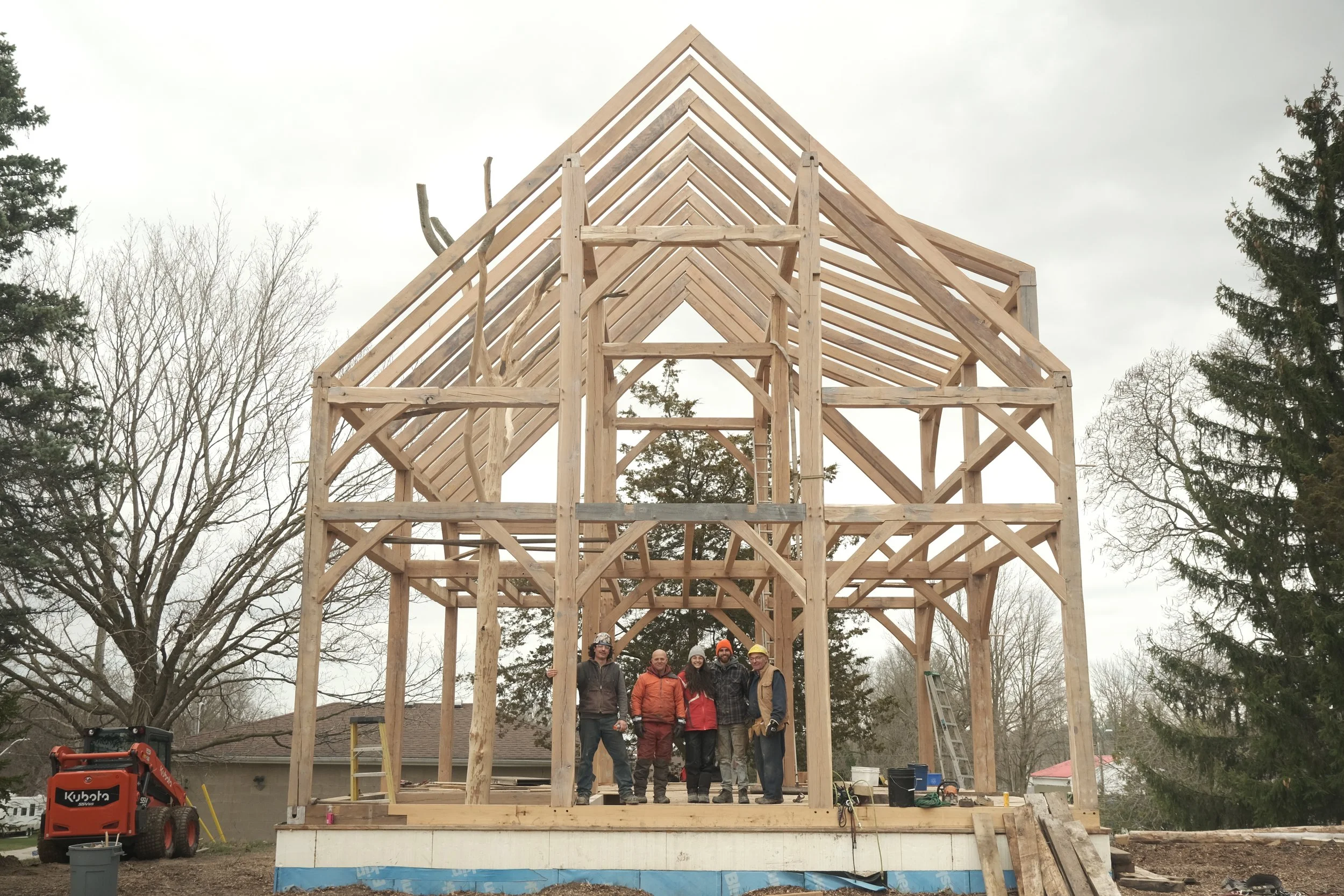Wooden house frame under construction with exposed beams and roof support, with some trees and neighboring houses in the background.