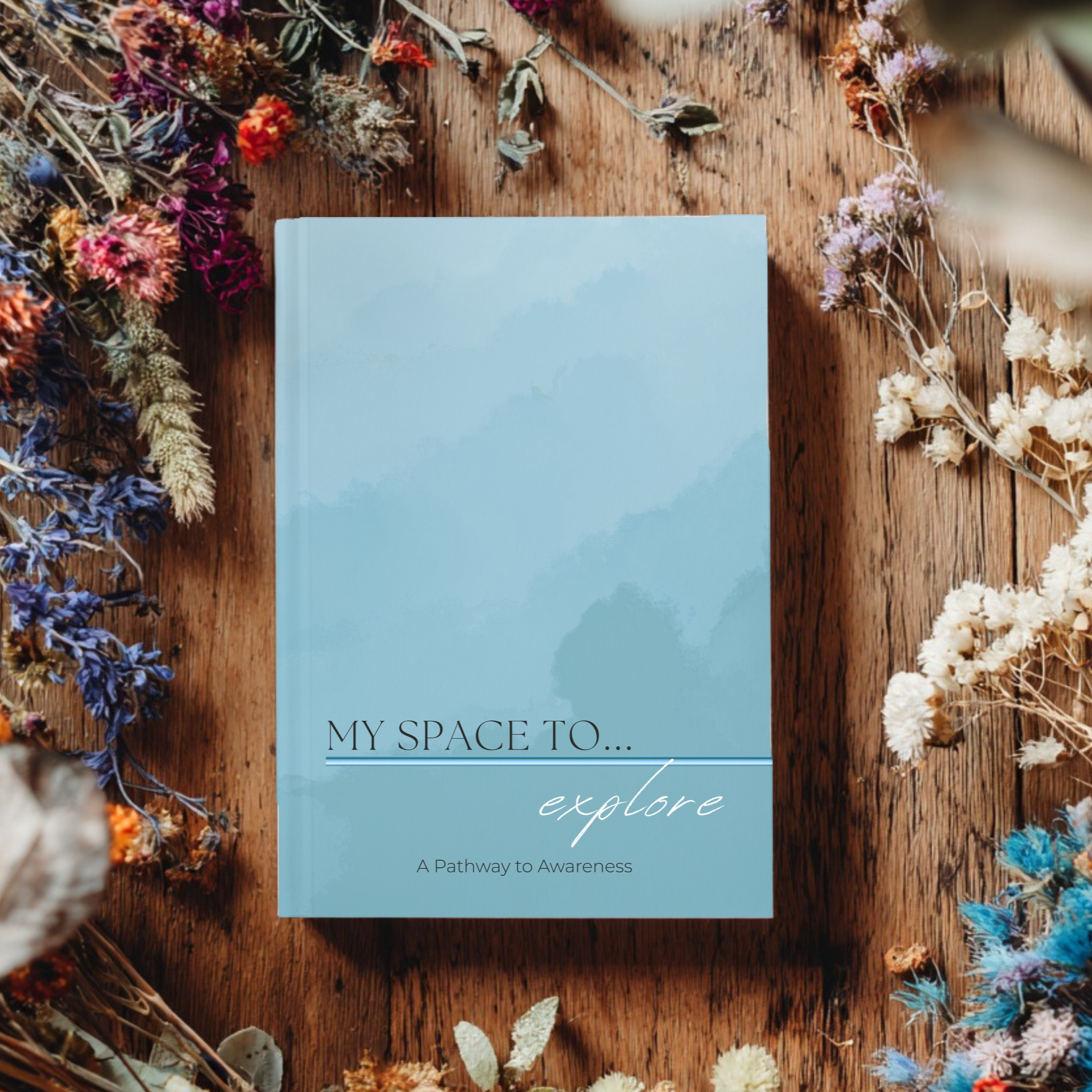 A light blue hardcover book titled 'My Space To... explore' with a subtitle 'A Pathway to Awareness,' on a wooden surface surrounded by colorful dried flowers.