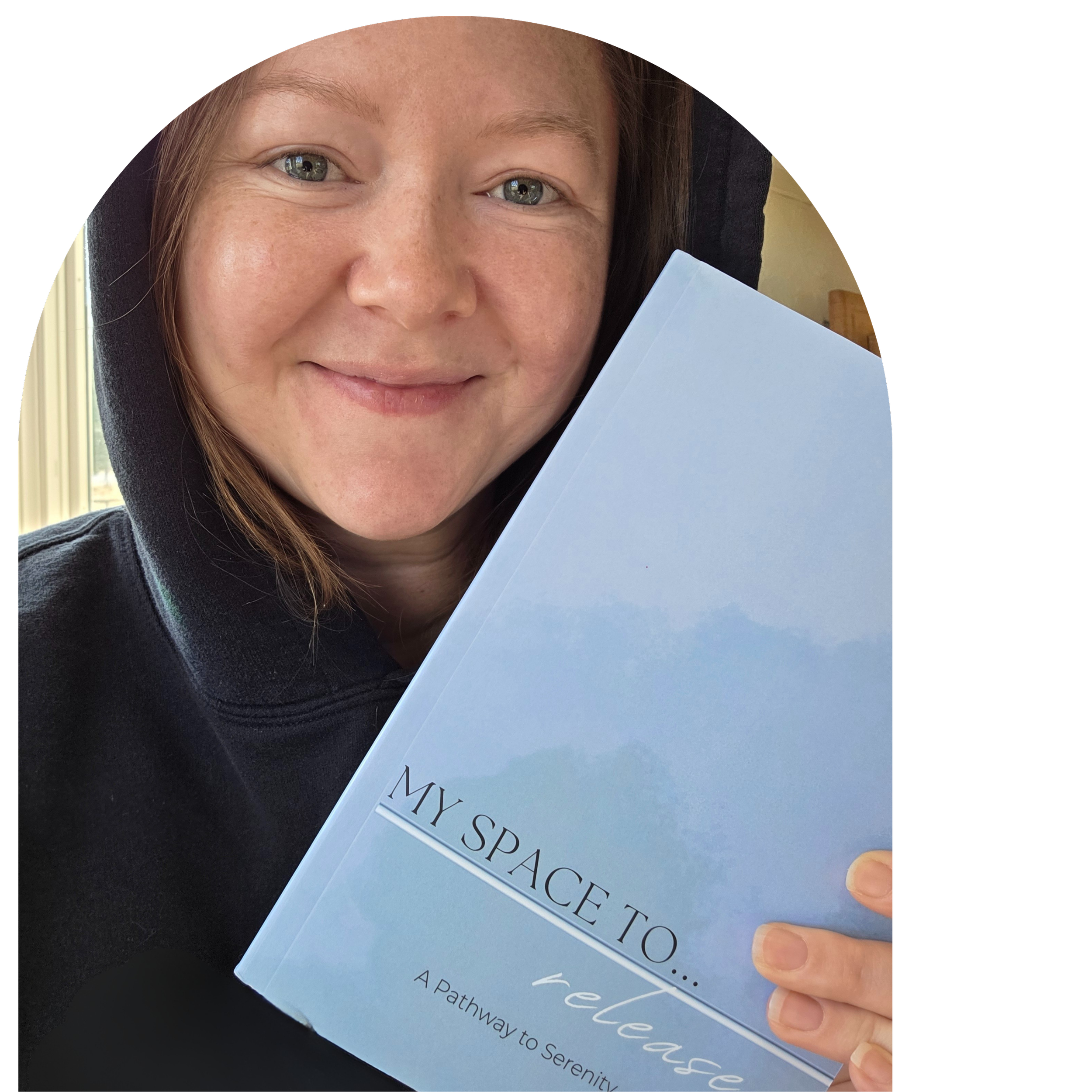 A woman smiling while holding a light blue book titled "My Space To..." with the subtitle "release" and "A Pathway to Serenity" underneath. She wears a black hoodie and has light brown hair, sitting indoors near a window.