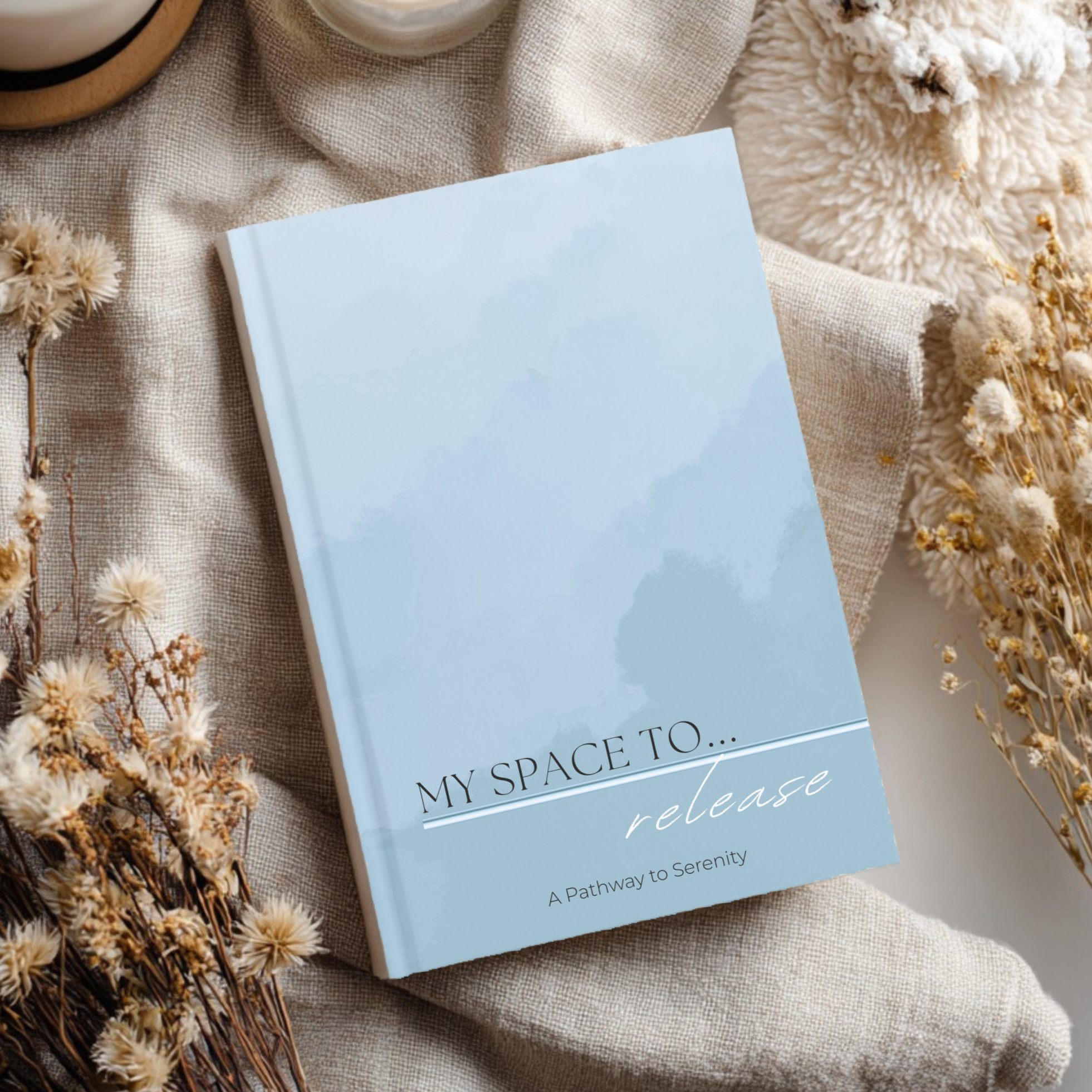 A light blue book titled "My Space to... release" with a subtitle "A Pathway to Serenity," surrounded by dried flowers and beige fabric.