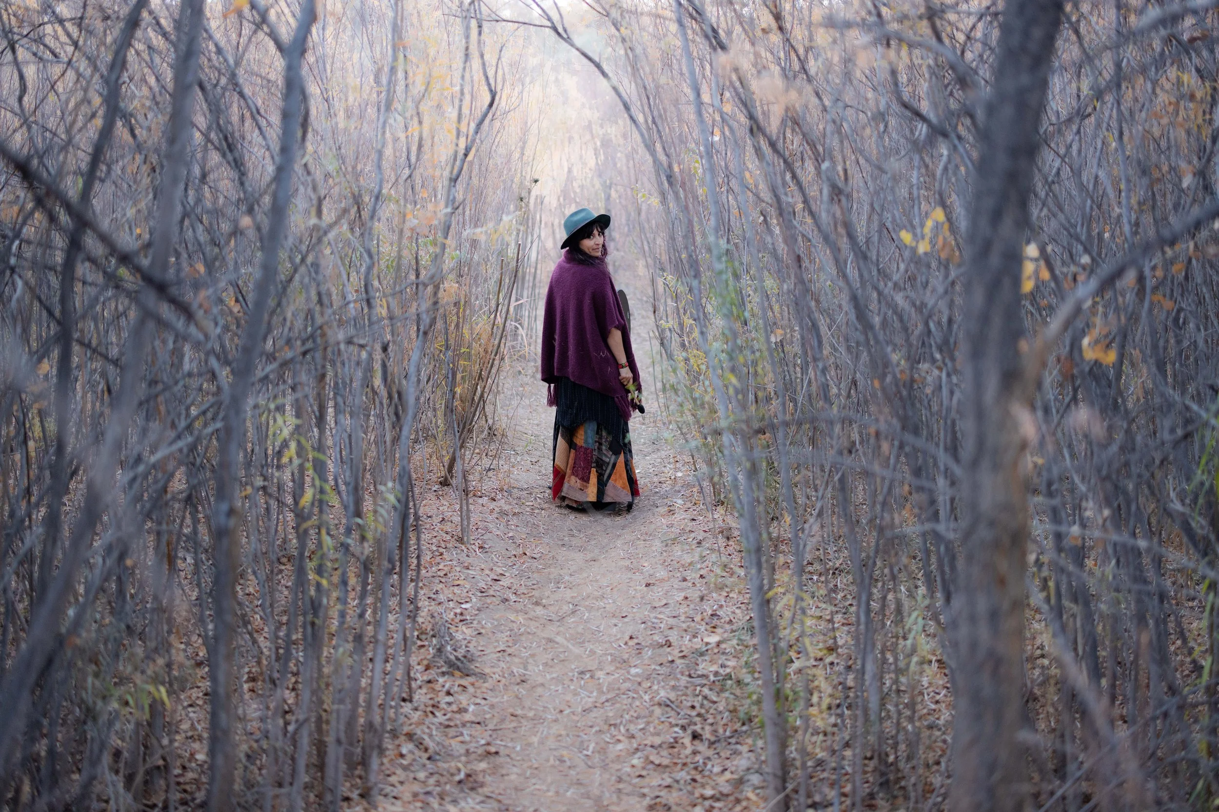 Sex, Relationship and life coach Zhenya Novareign standing on a dirt path in a forest with leafless trees and fallen leaves, wearing a wide-brimmed hat, a purple shawl, and a colorful patchwork skirt.