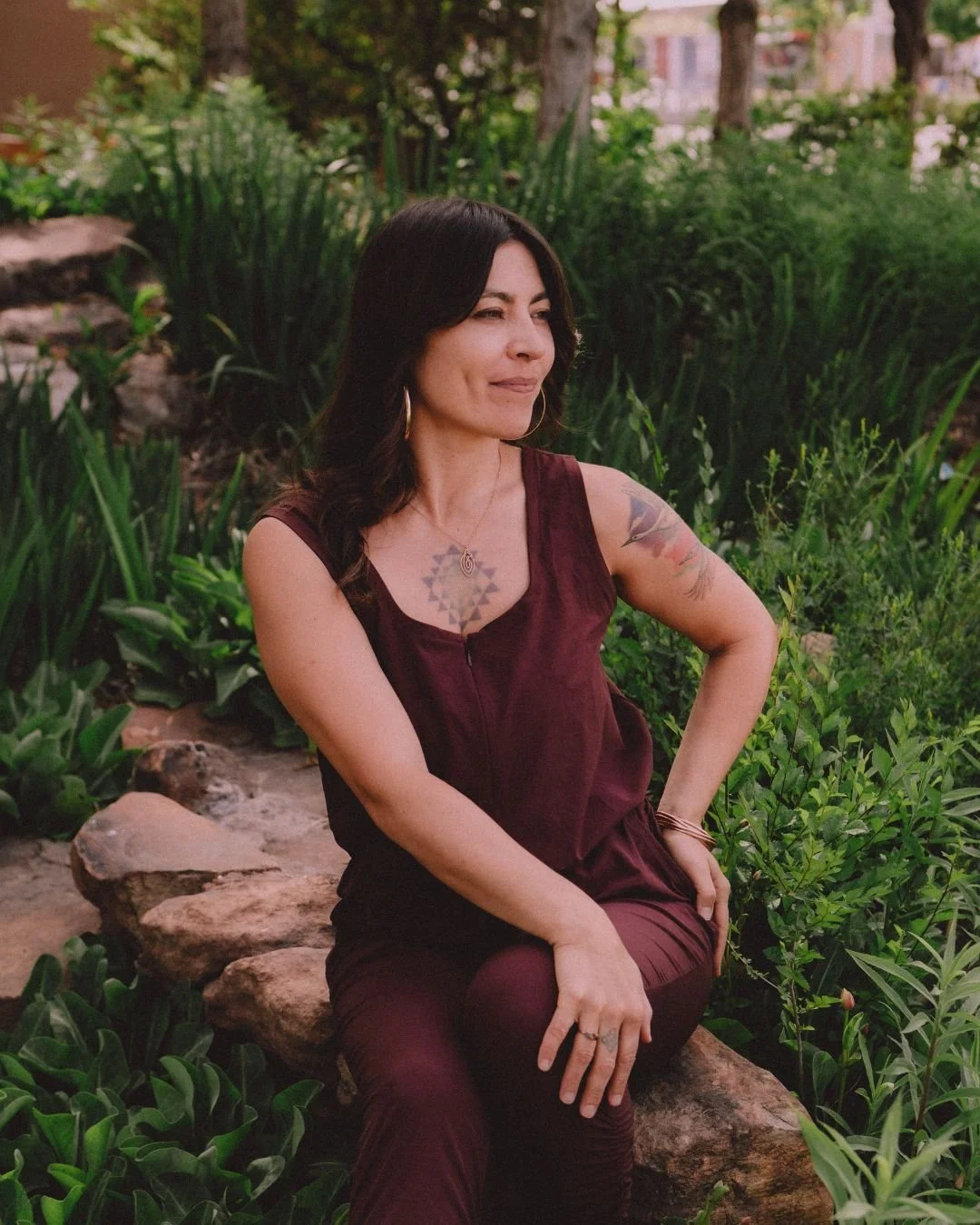 A woman with dark hair and tattoos on her arms sitting on a rock in a lush garden with green plants.