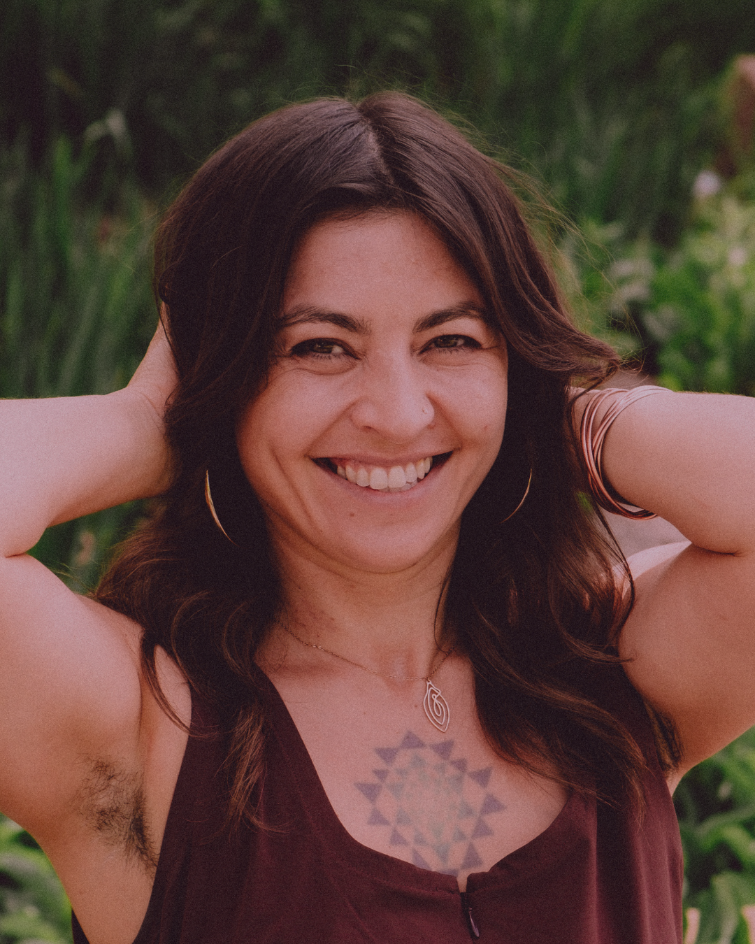 A woman smiling outdoors, with arms raised behind her head, wearing hoop earrings, a necklace, and a sleeveless top, with a tattoo on her chest, on a green background.