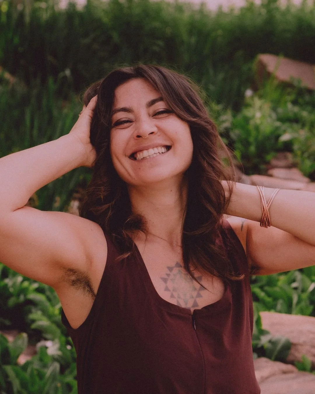 A smiling woman with wavy brown hair, tattoos, and a sleeveless top standing outdoors among greenery.
