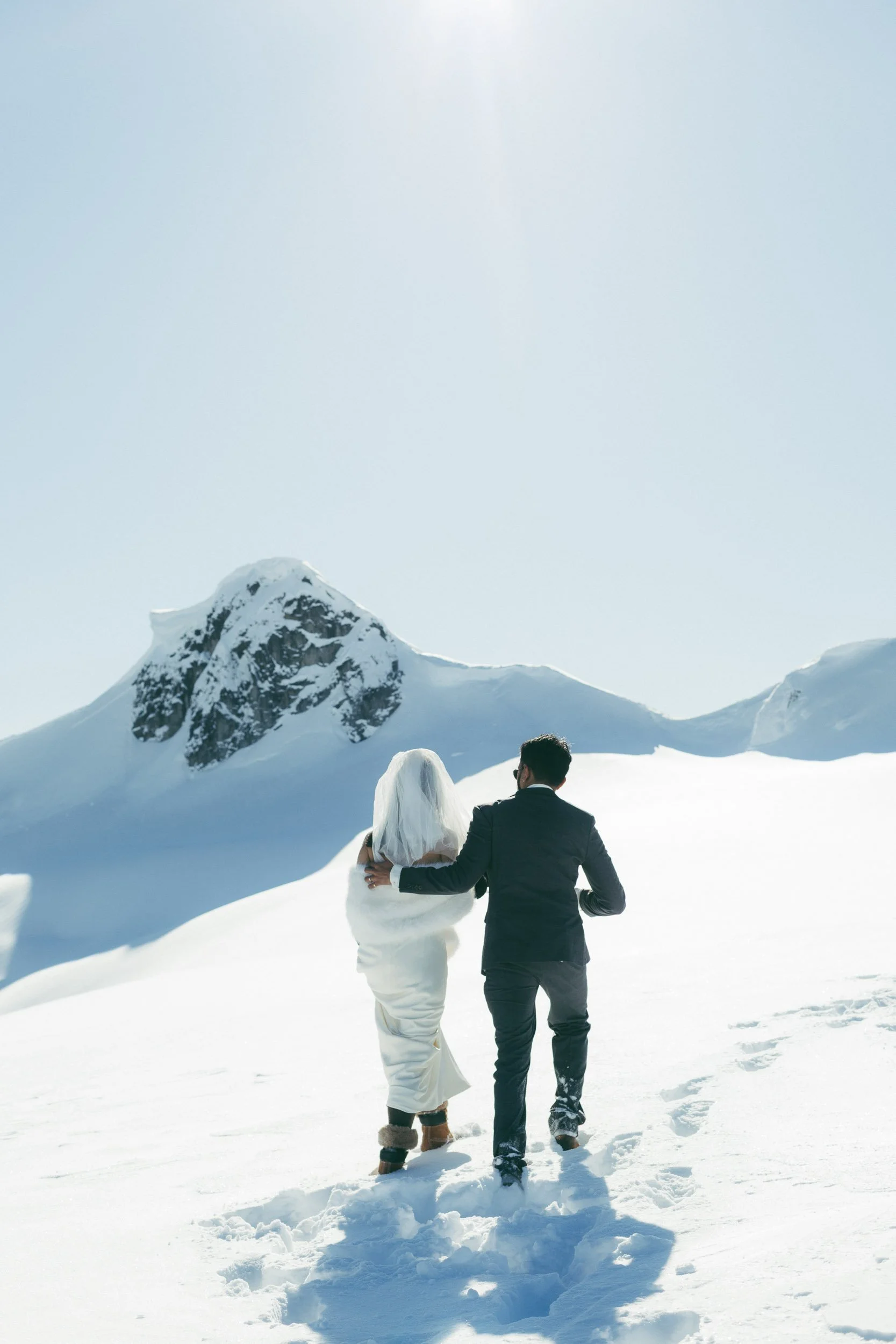 A bride and groom walking in the snow in a mountain landscape with snow-covered peaks under a clear blue sky.