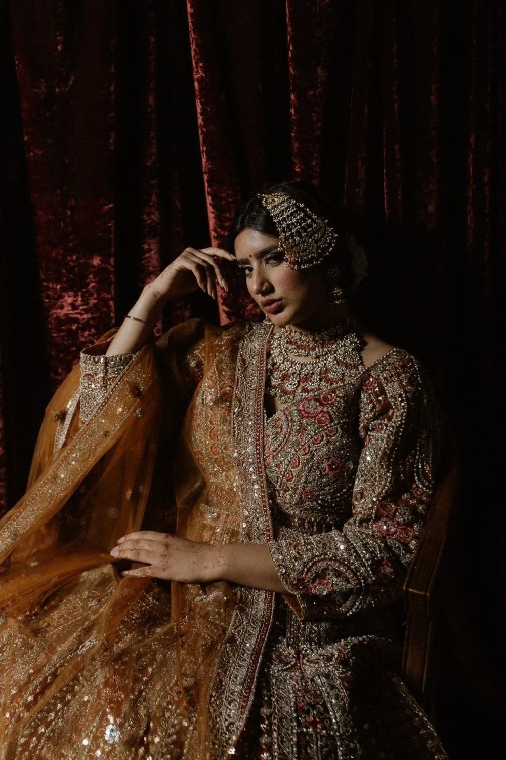 A South Asian bride in an embroidered lehenga seated before red velvet curtains