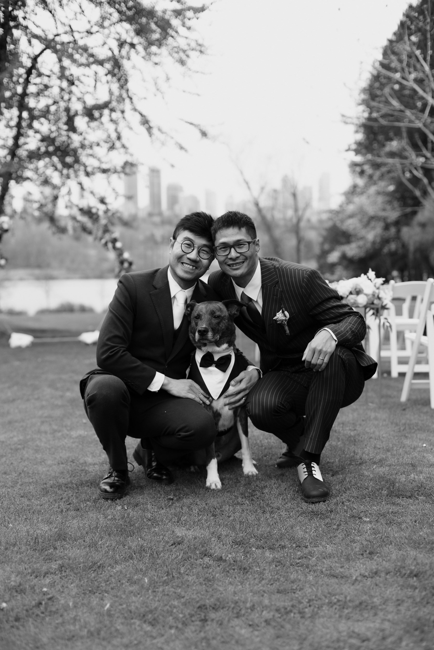 Two men in suits with glasses and a dog in a tuxedo; they are outdoors on a lawn during daytime, smiling and kneeling close together, with city buildings in the blurry background.