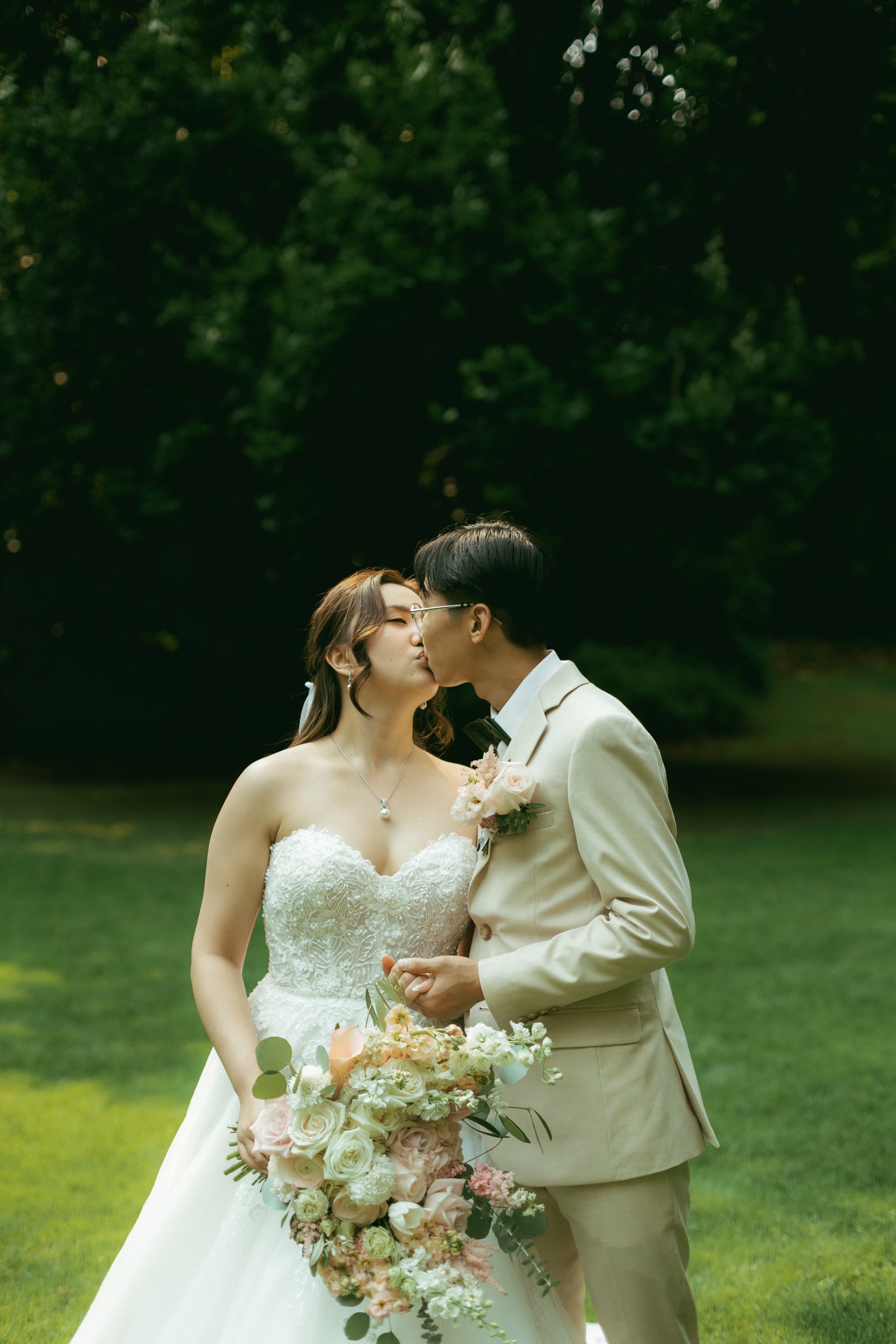 A bride and groom kissing outdoors in a lush green park with trees in the background. The bride wears a strapless white wedding dress and holds a large bouquet of flowers, while the groom is dressed in a cream-colored suit with glasses.
