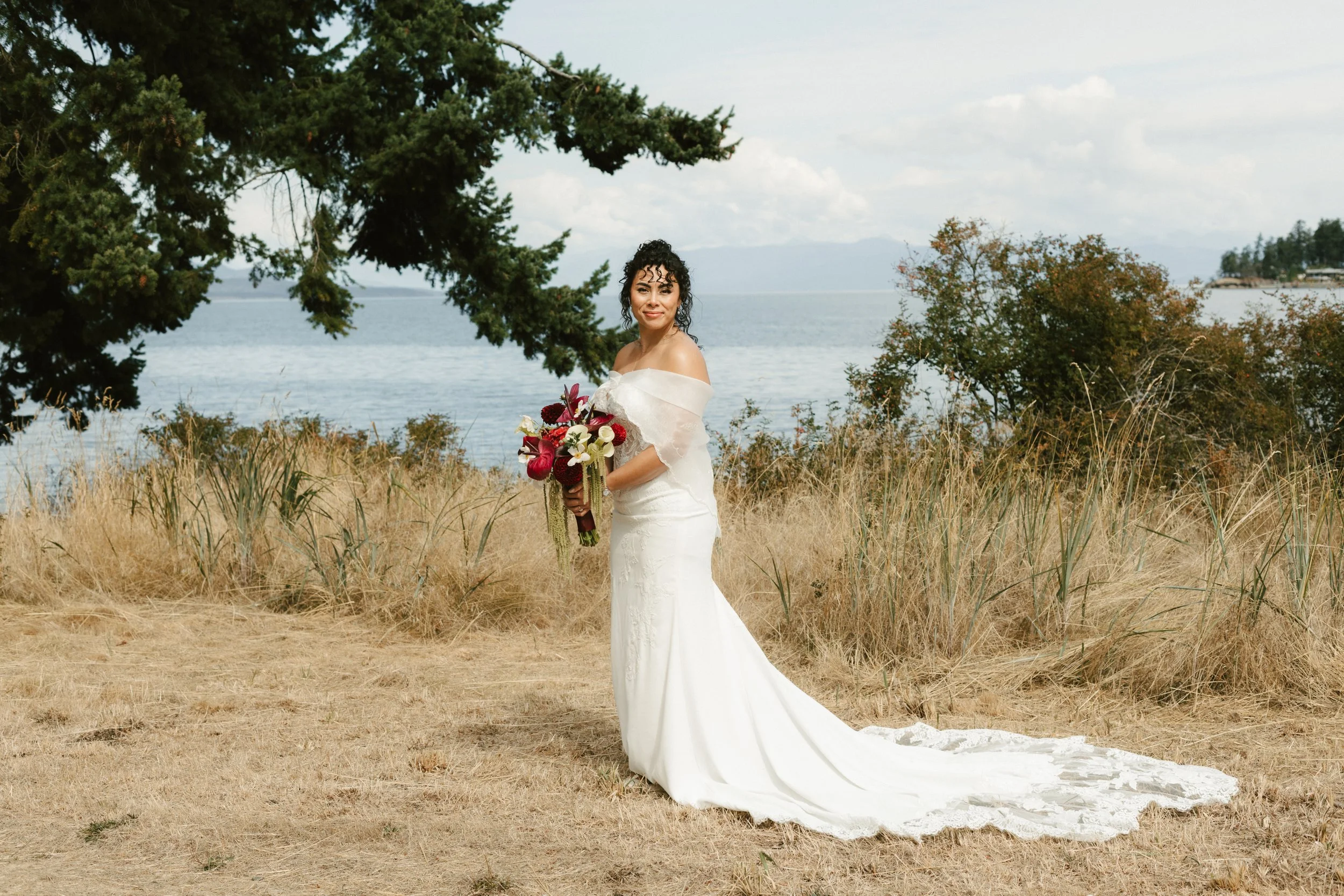 Bride in a white wedding gown holding a colorful bouquet beside a lake in a grassy field