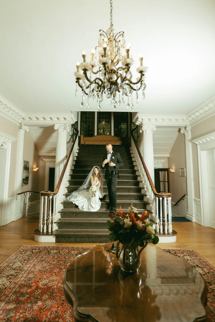 A couple on a grand staircase beneath a crystal chandelier at Hycroft Manor