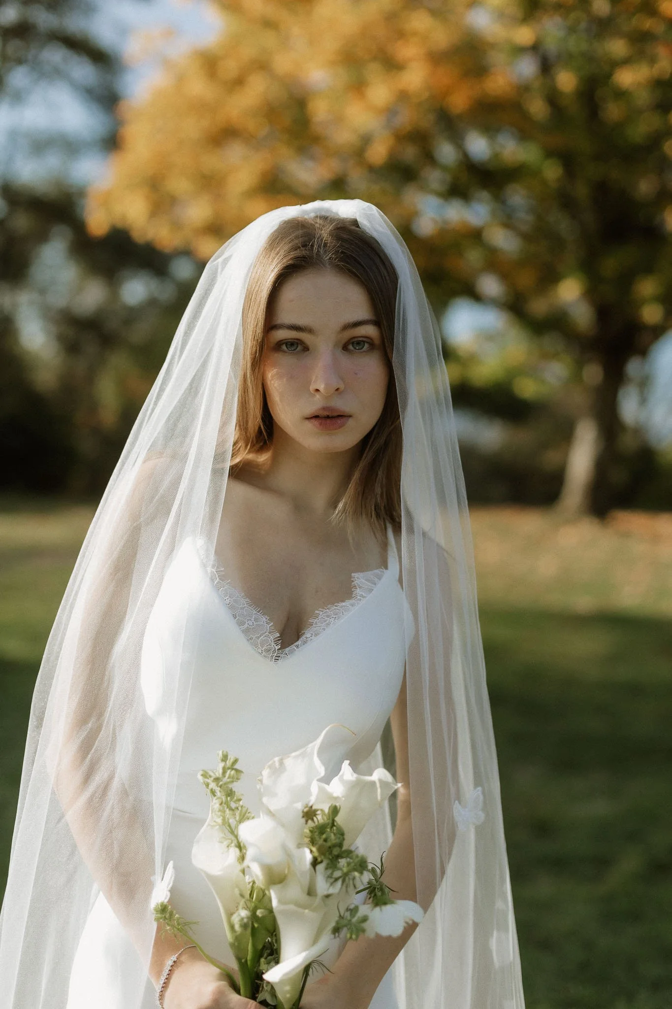A bride in a long veil holding white calla lilies beneath an autumn tree