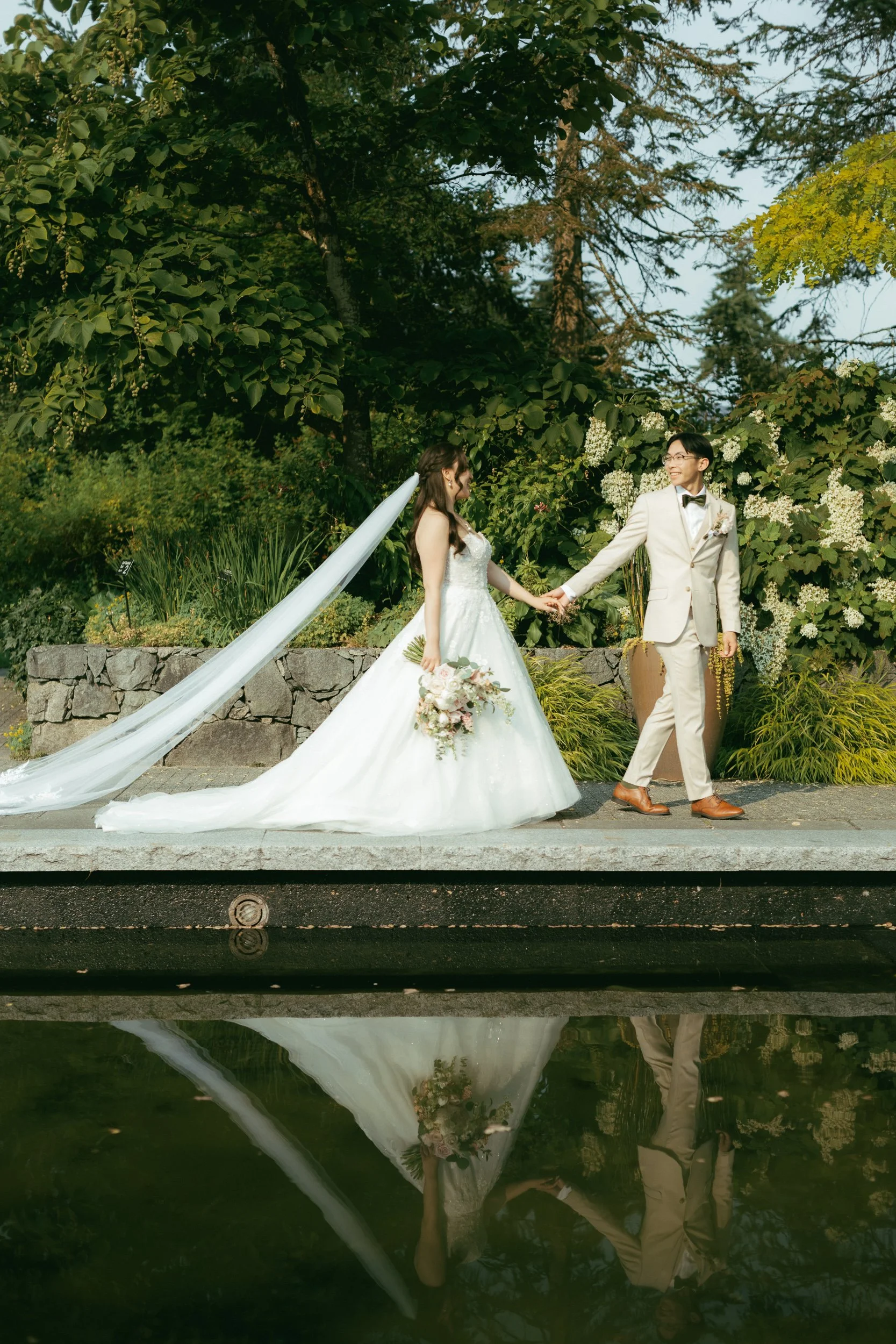 A newlywed couple holding hands and smiling outdoors in a garden setting, with lush green trees and plants around them, their reflection visible in a nearby body of water.