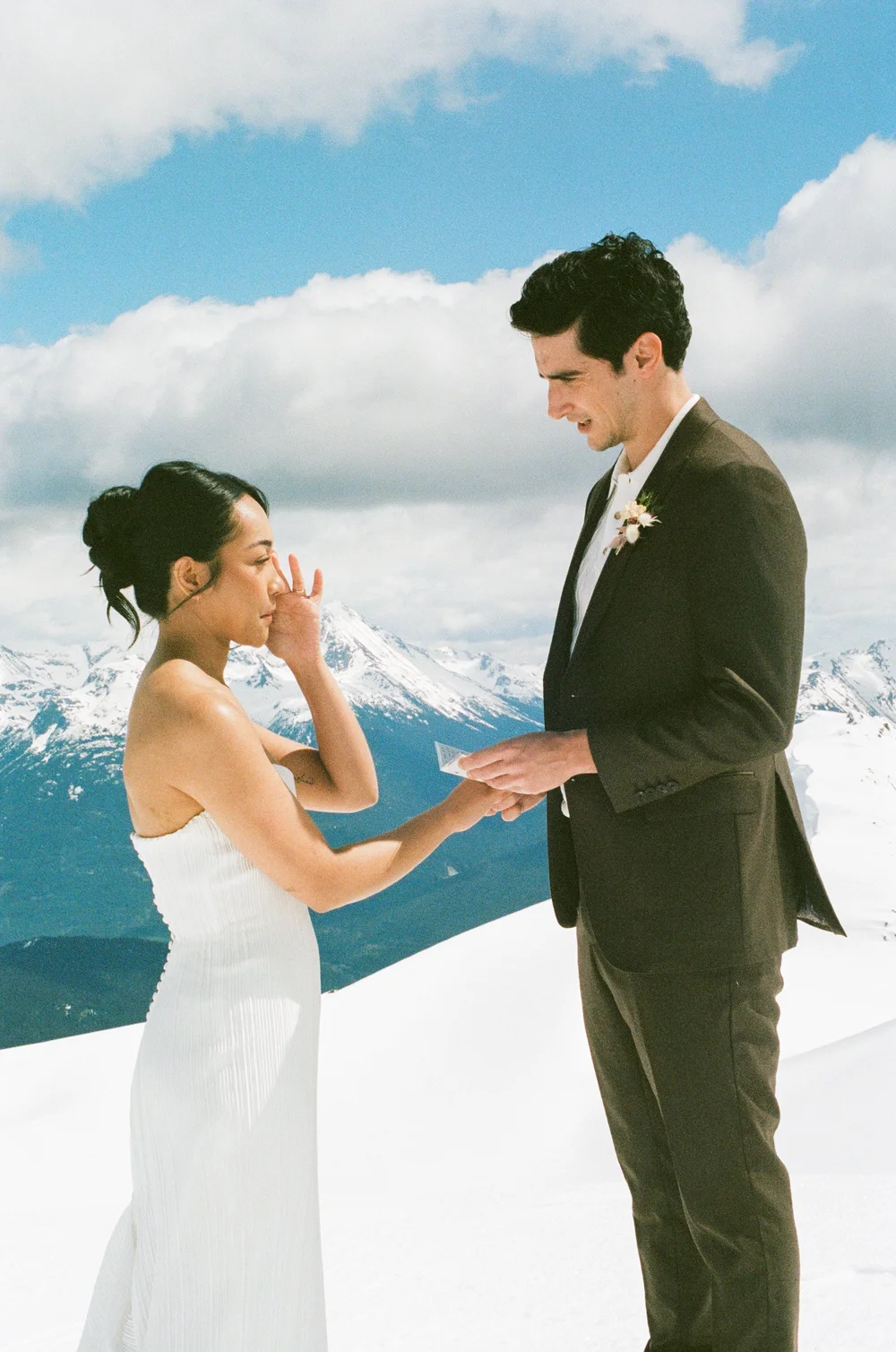 A bride wiping away a tear as her groom holds her hands on a snowy summit, photographed on 35mm film
