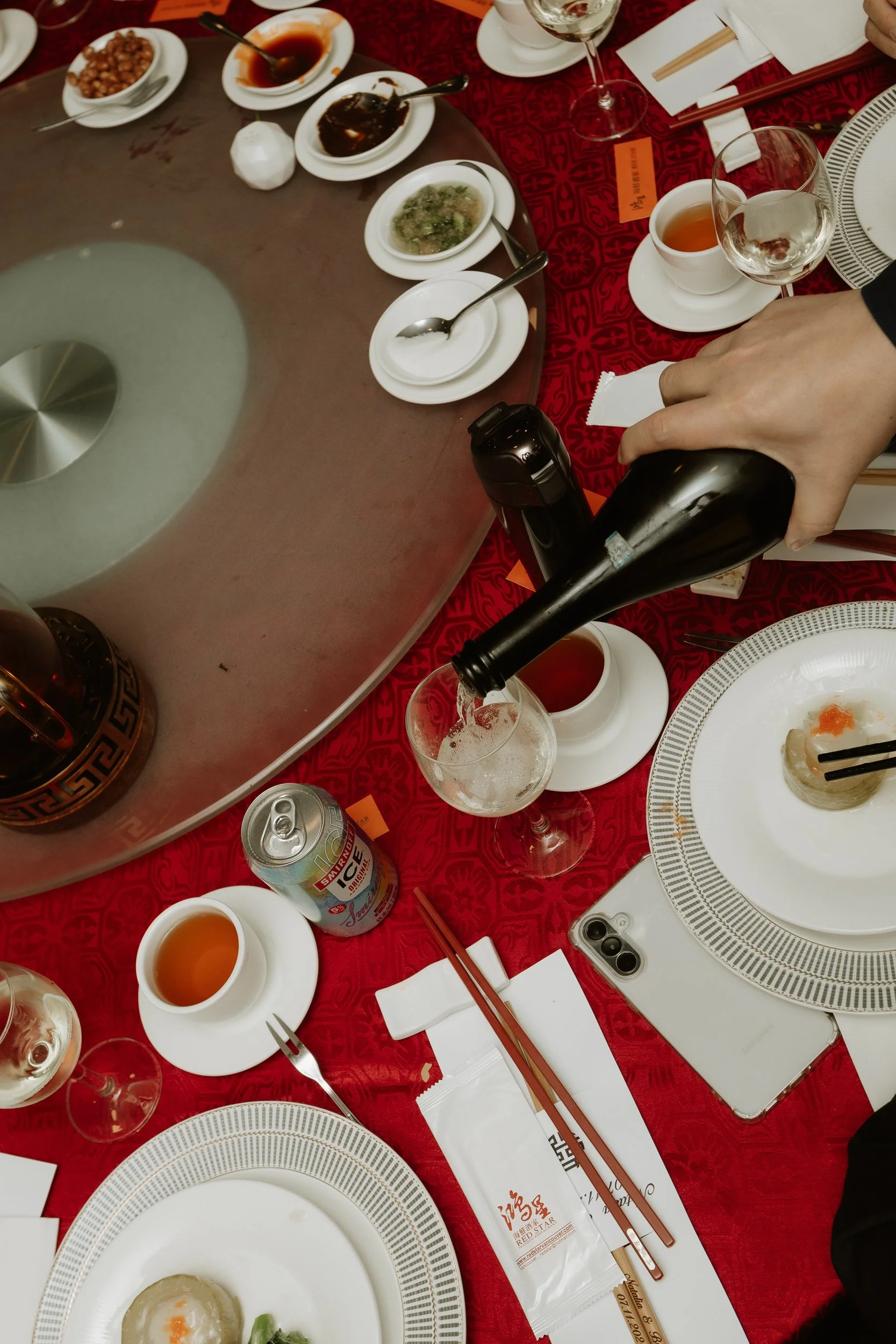 A table set for a meal with plates, cups, wine glasses, chopsticks, and various dishes including a dish topped with an egg, bowls of sauce, and a glass of beer. A person's hand is pouring a drink into a wine glass.
