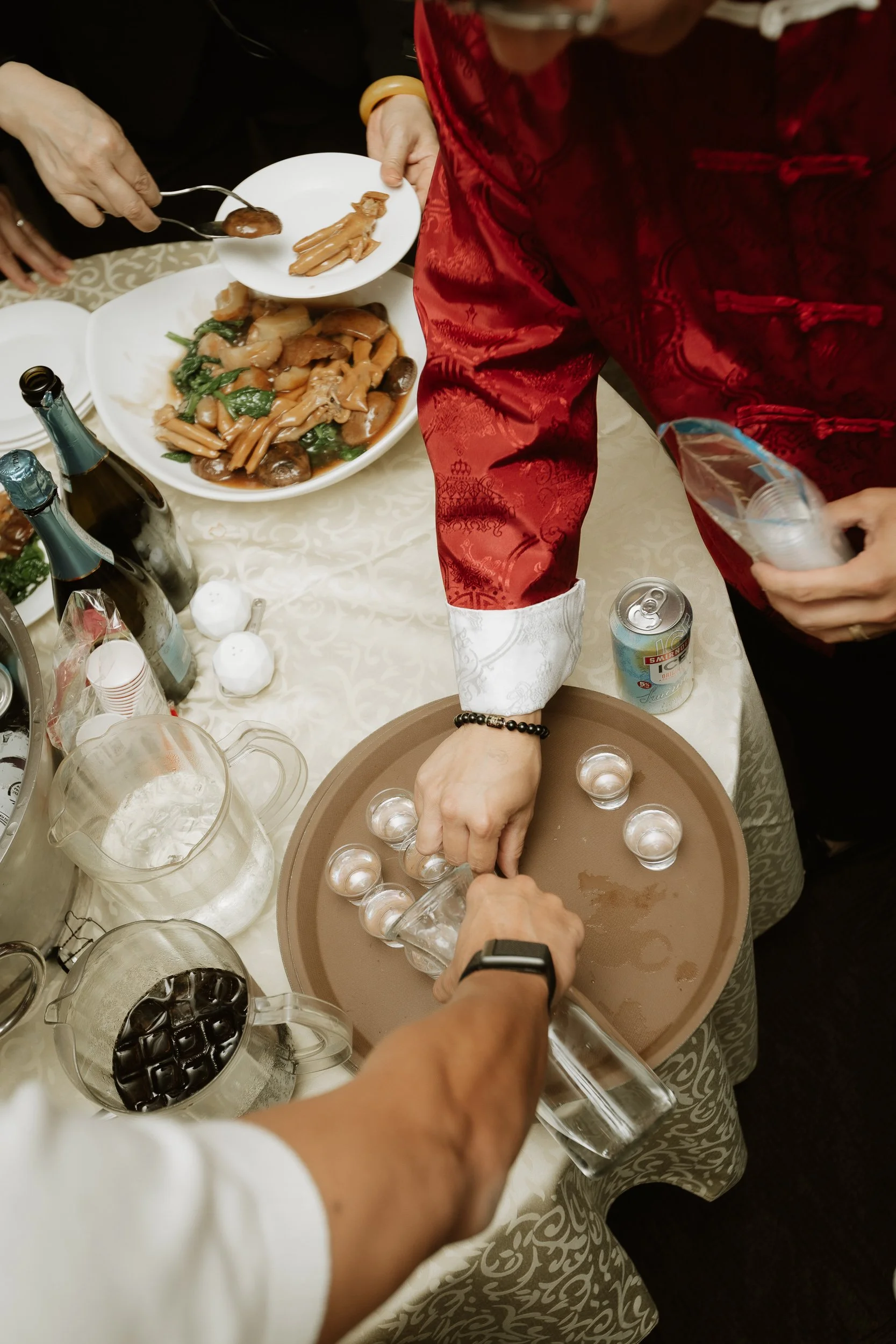 People at a table preparing and serving drinks and food, with dishes, bottles, and glasses visible on the table.