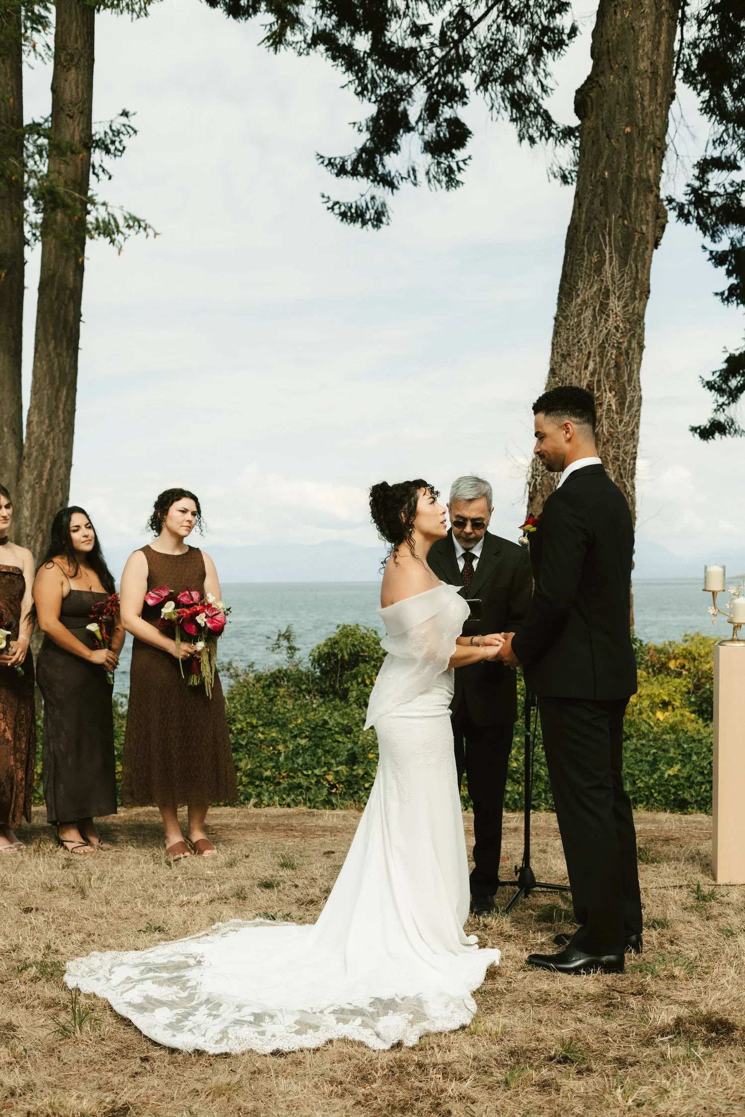 A bride and groom exchanging vows beneath tall pines, the sea behind them