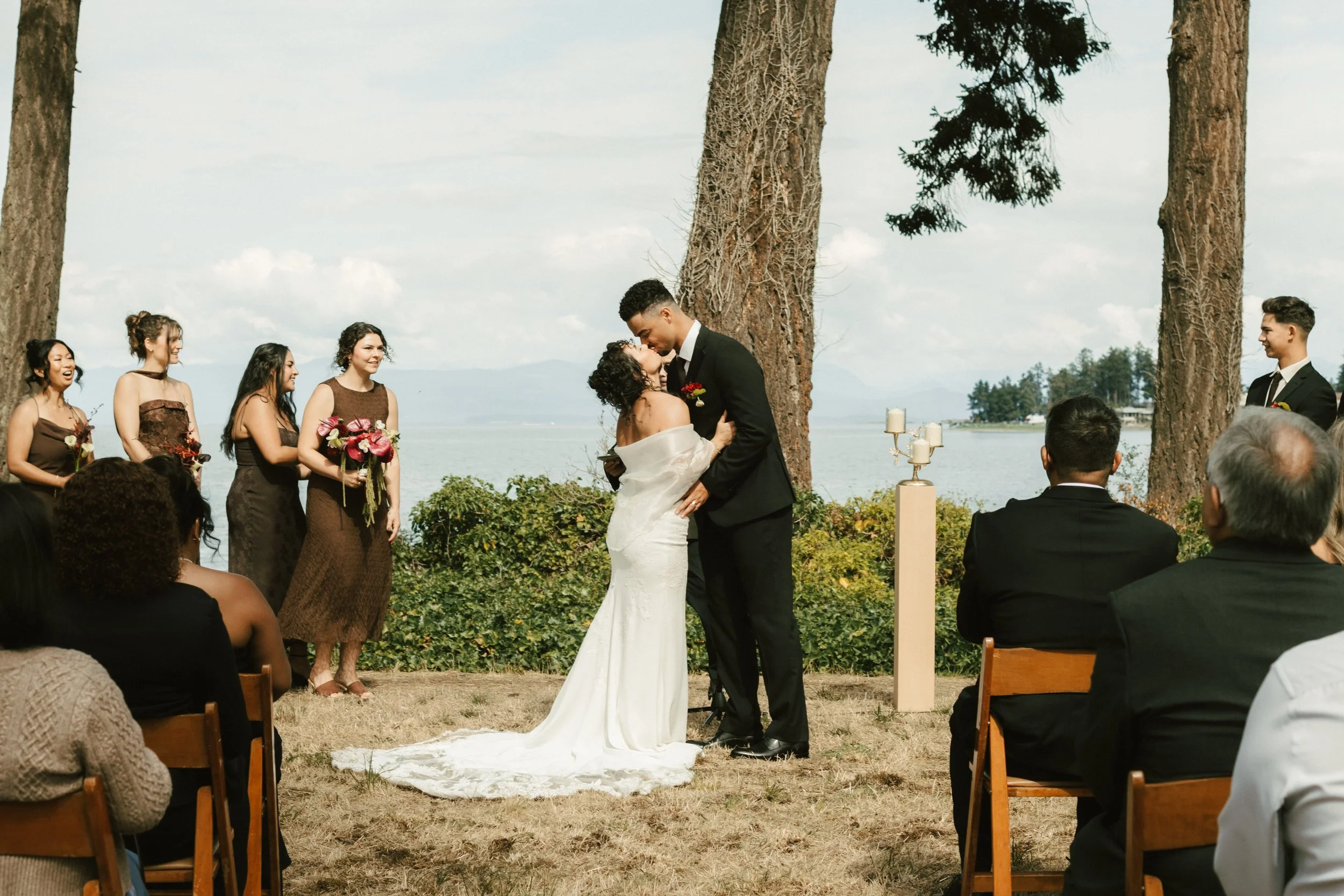 Bride and groom kiss during an outdoor lakeside wedding ceremony surrounded by attendants and seated guests