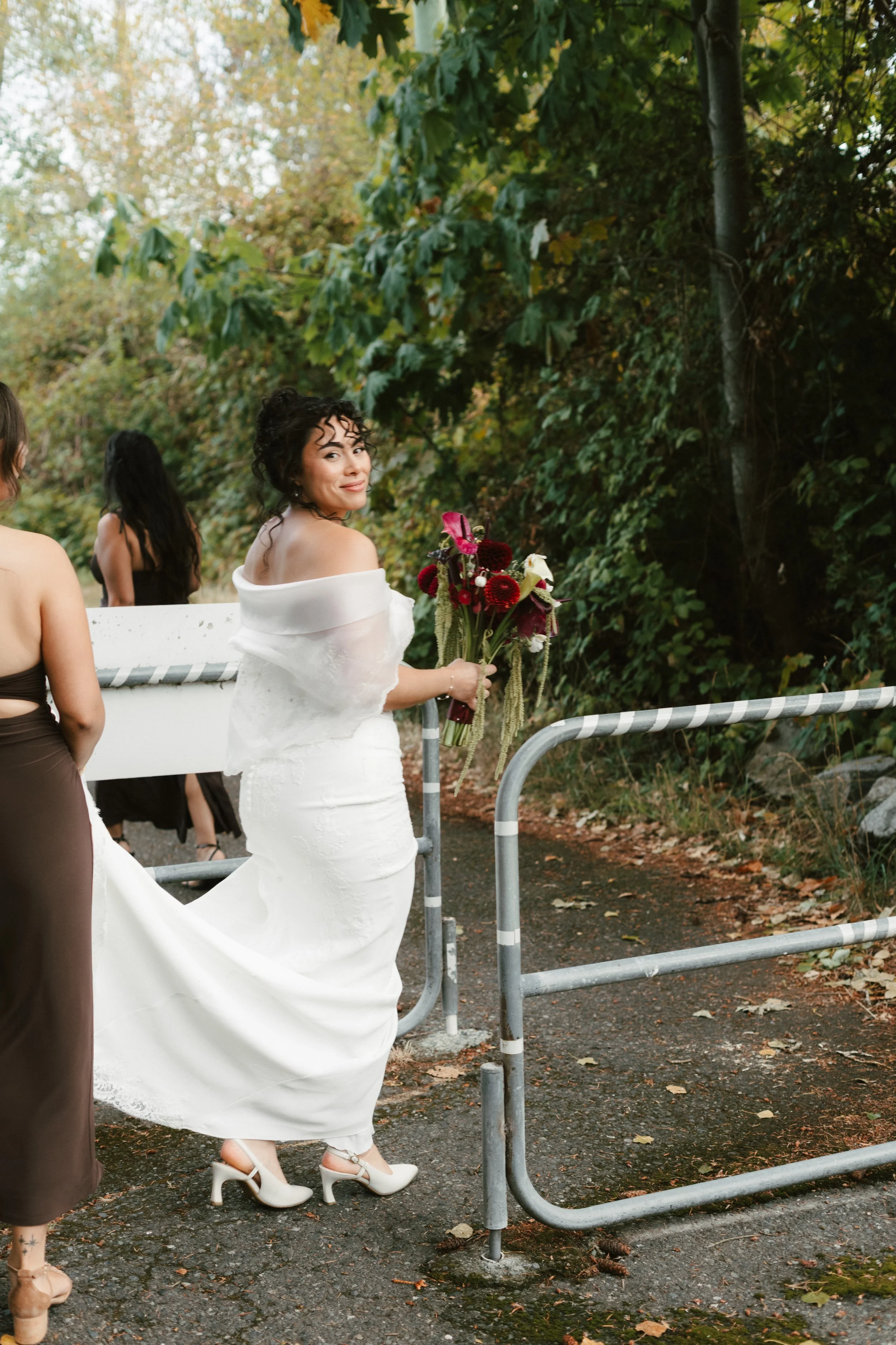 Bride in a white wedding dress holding a colorful bouquet outdoors with bridesmaids in a leafy forest setting