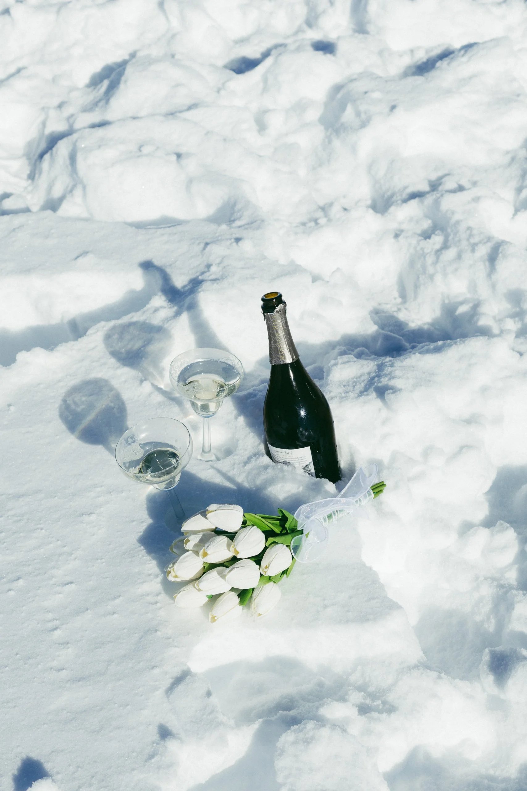 Bottle of champagne, two glasses of champagne, and a bouquet of white tulips on snow