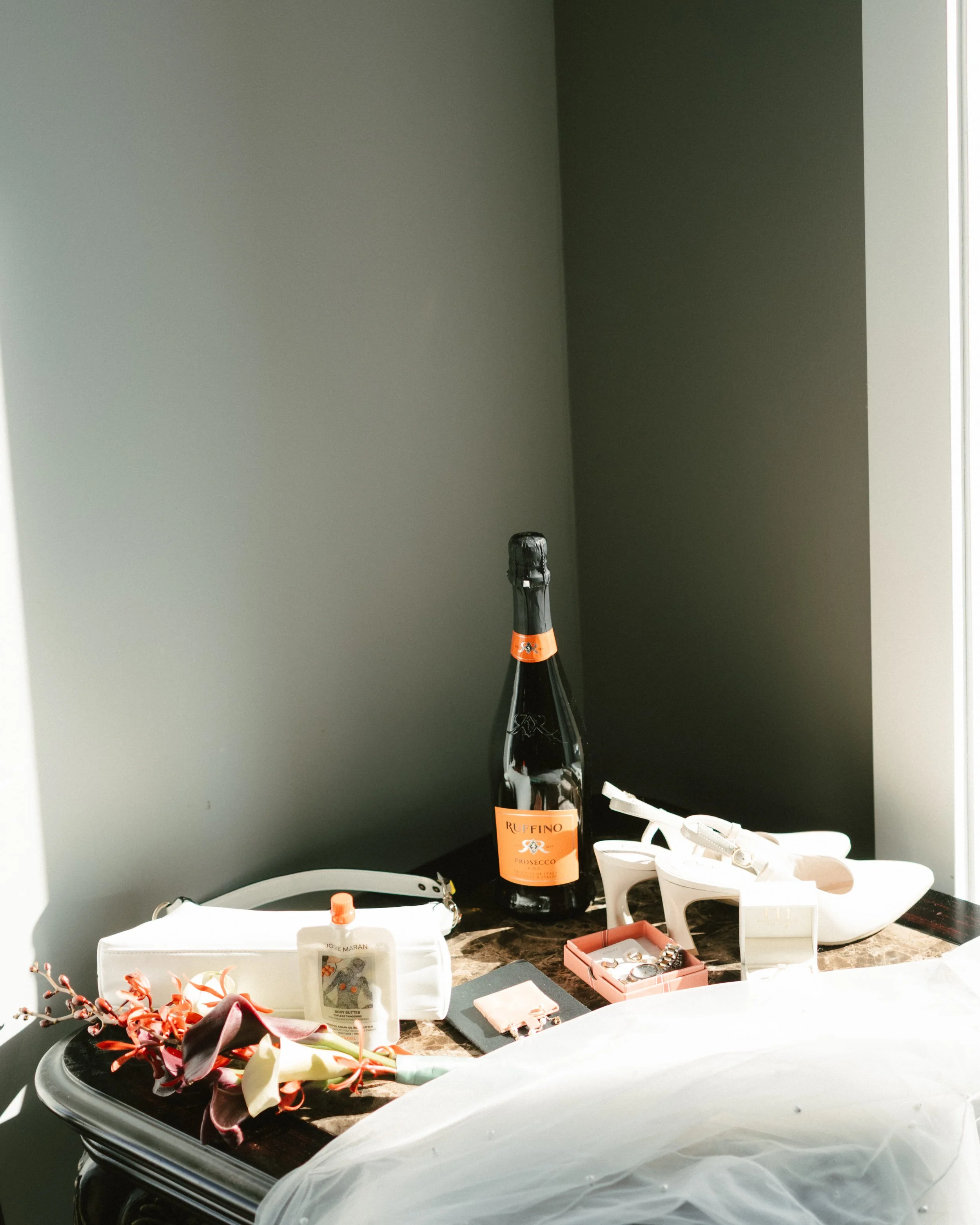 Ruffino Prosecco bottle surrounded by white bridal shoes, jewelry, body butter, flowers, and wedding accessories on a marble vanity