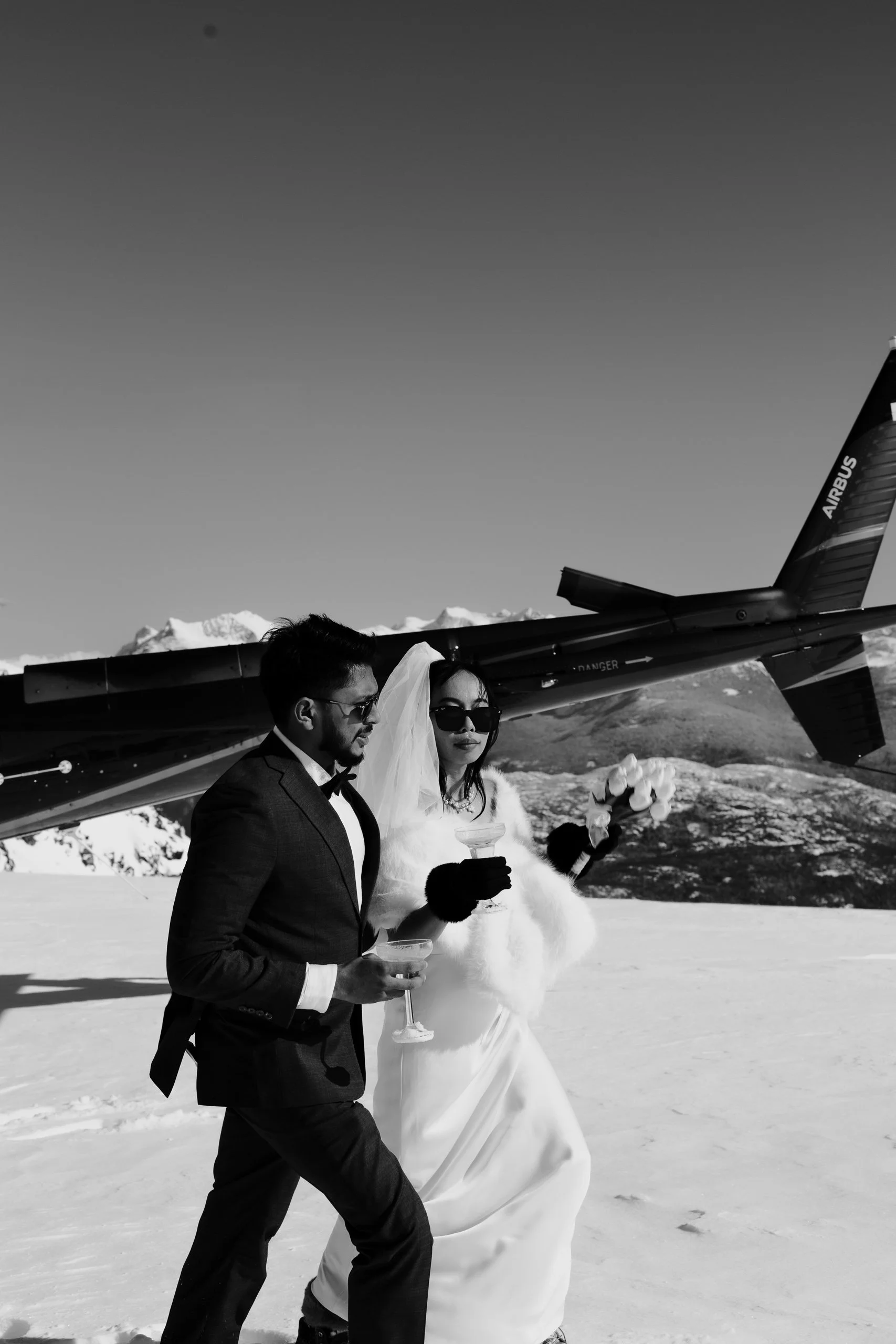 A man in a suit and a woman in a wedding dress wearing sunglasses, holding drinks and flowers, walking in an icy landscape with snow-capped mountains and a helicopter overhead.
