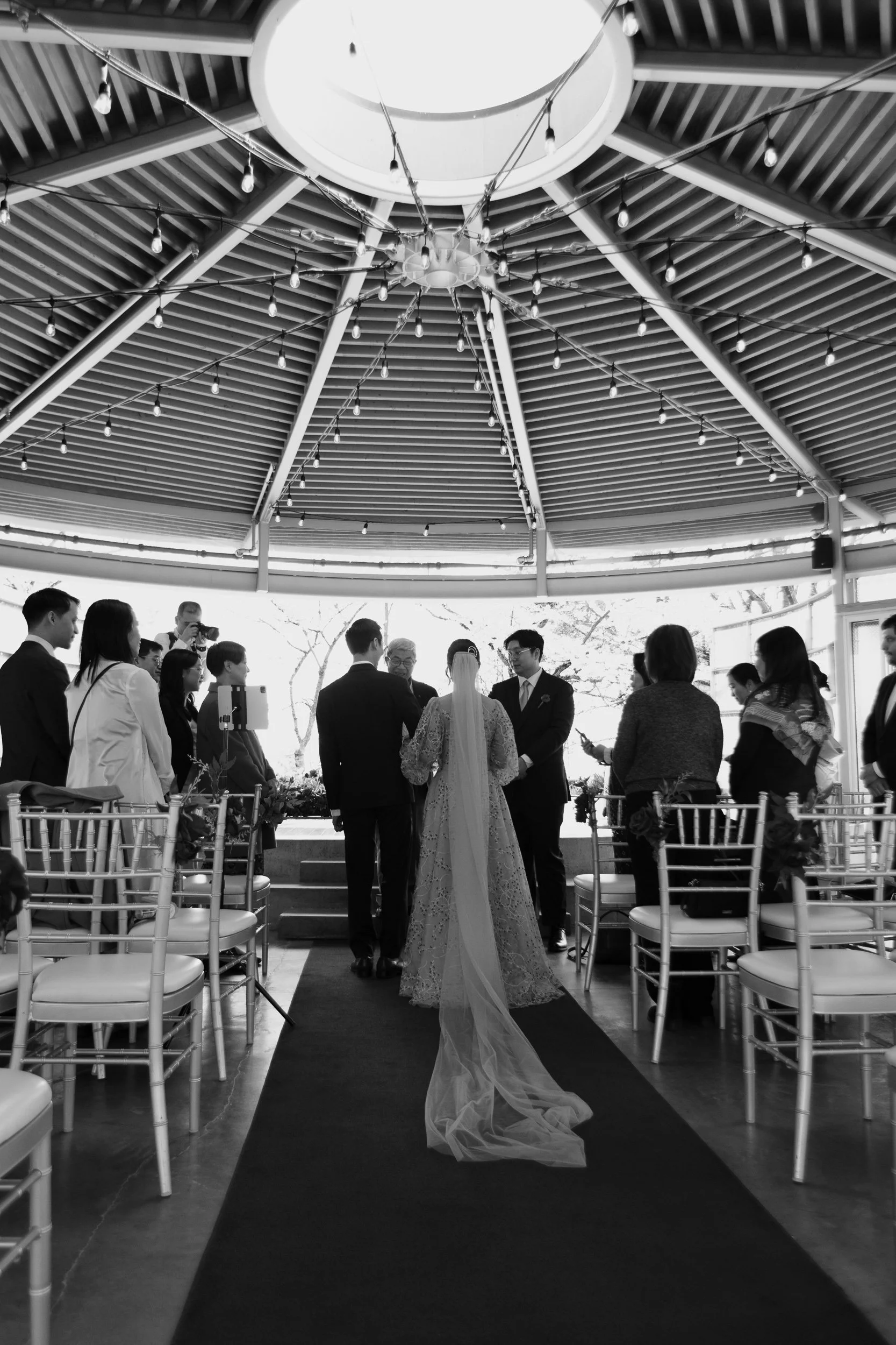 Black and white photo of a wedding ceremony with a bride and groom standing at the altar, surrounded by guests, in a decorated outdoor or semi-outdoor venue with a circular ceiling and string lights.