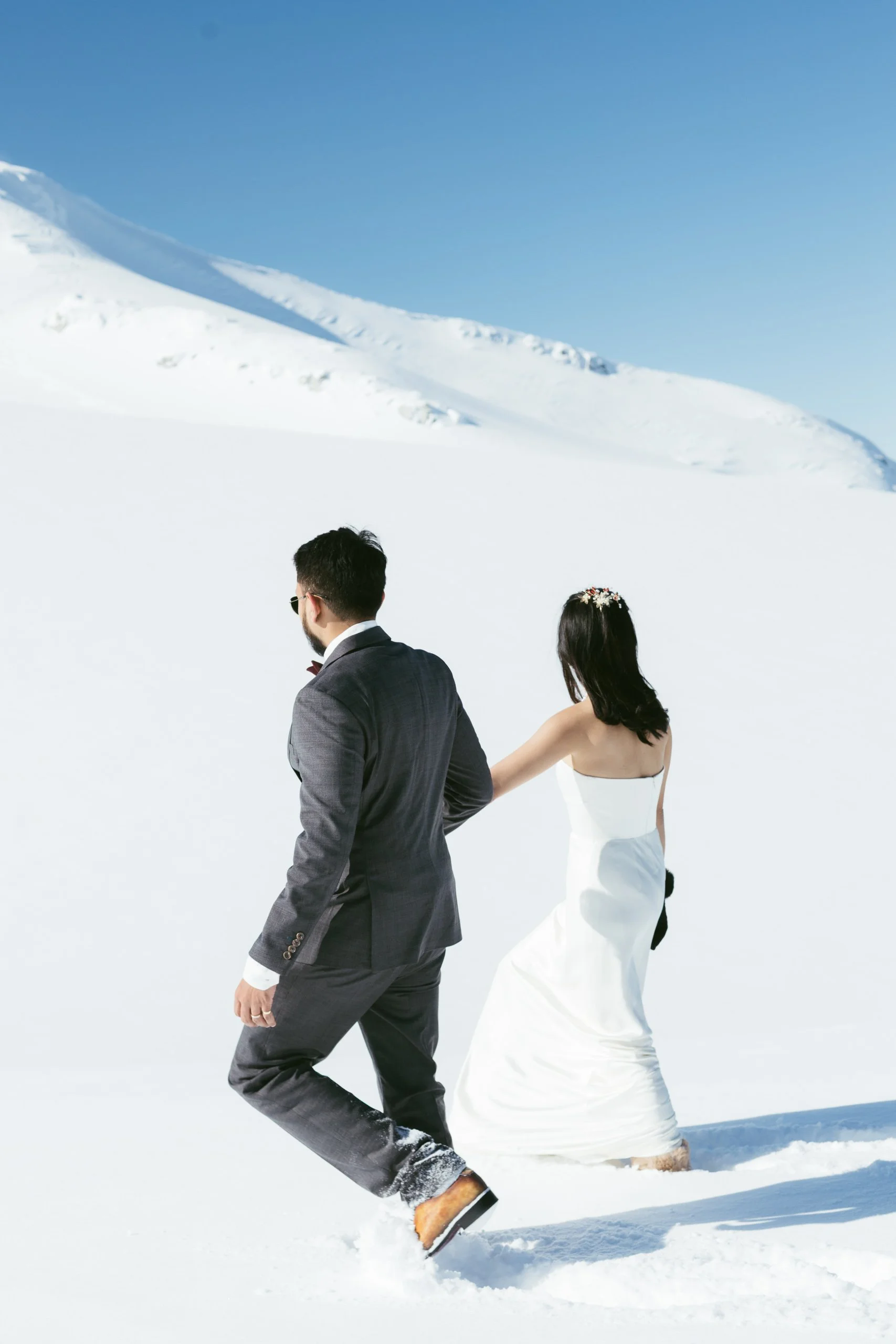 A couple dressed in wedding attire walking through snow-covered landscape with snow mountains in the background.