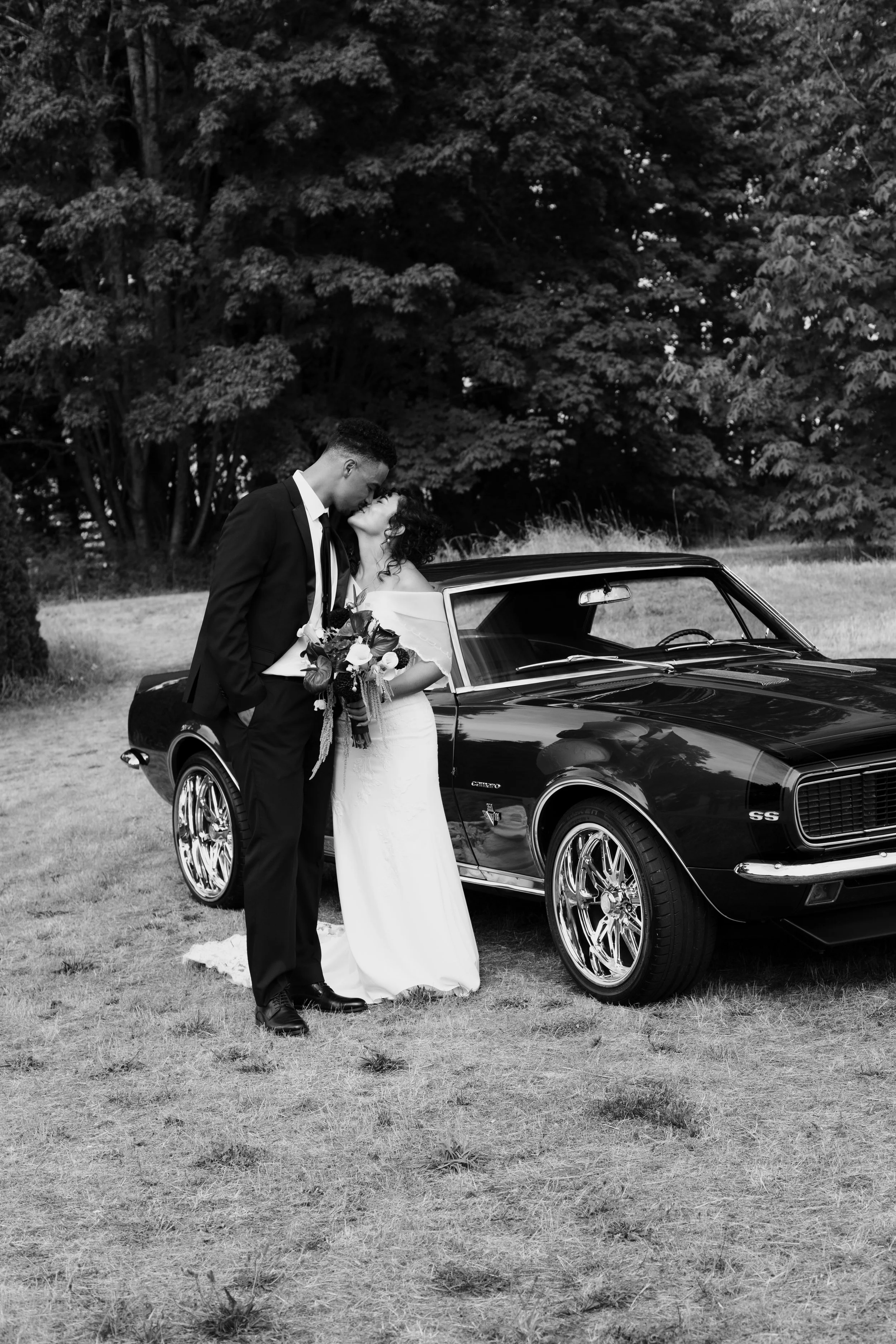 Newlywed couple kissing beside a classic Chevrolet Camaro in a wooded outdoor setting