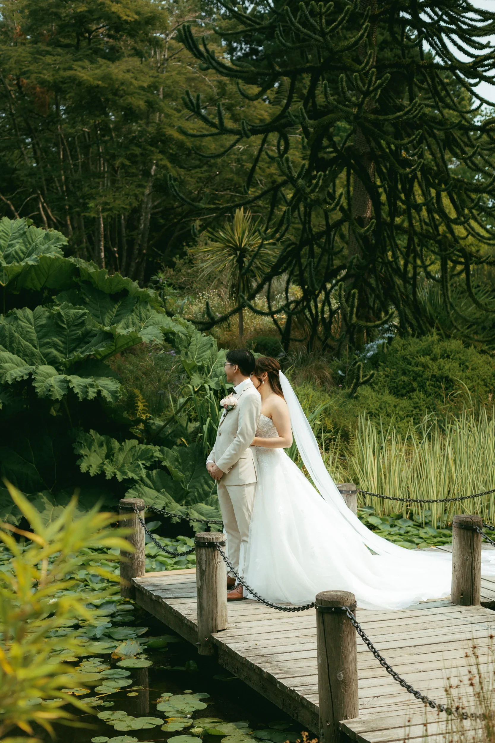 A bride and groom standing on a wooden bridge surrounded by lush greenery, with pond water and lily pads below.