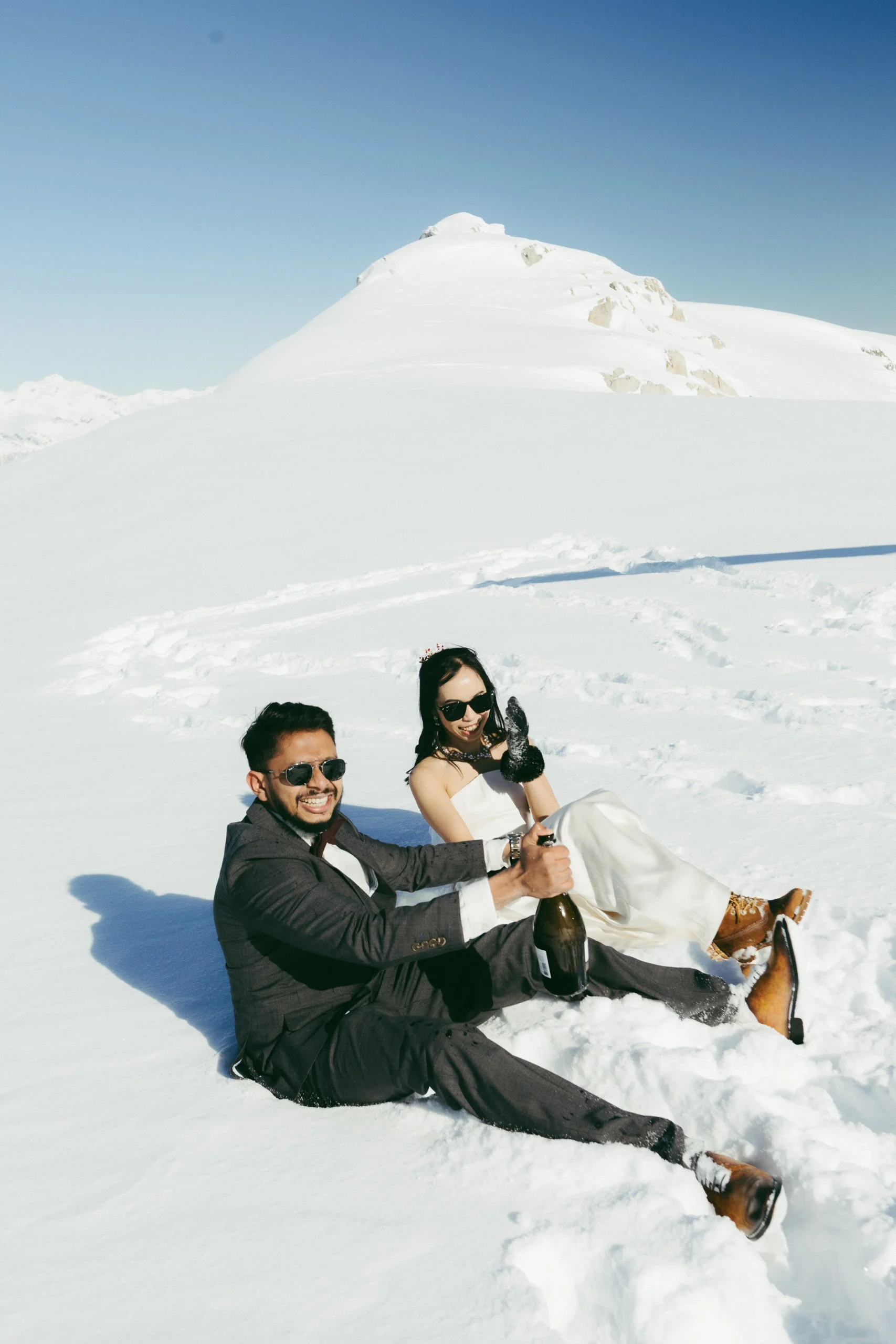 A man and woman sitting in the snow on a mountain, holding a champagne bottle, smiling, and wearing sunglasses and winter clothing