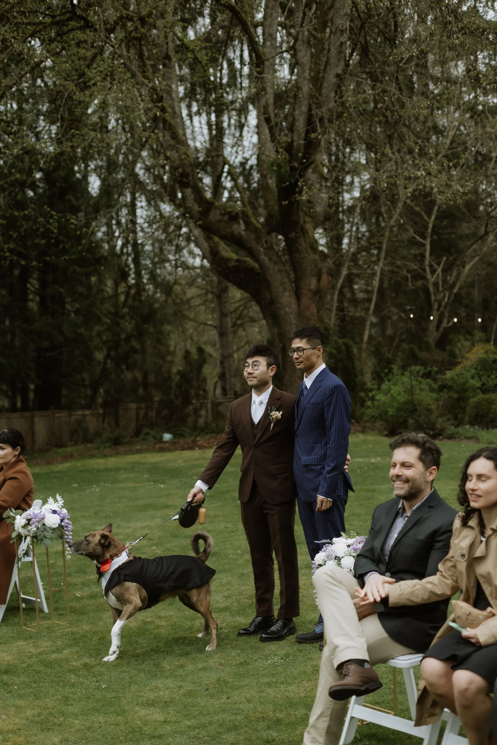 A group of people dressed in formal attire are attending an outdoor event in a park with large trees. Some individuals are seated, holding floral arrangements, while others are standing, including a man with a leashed dog wearing a tuxedo.