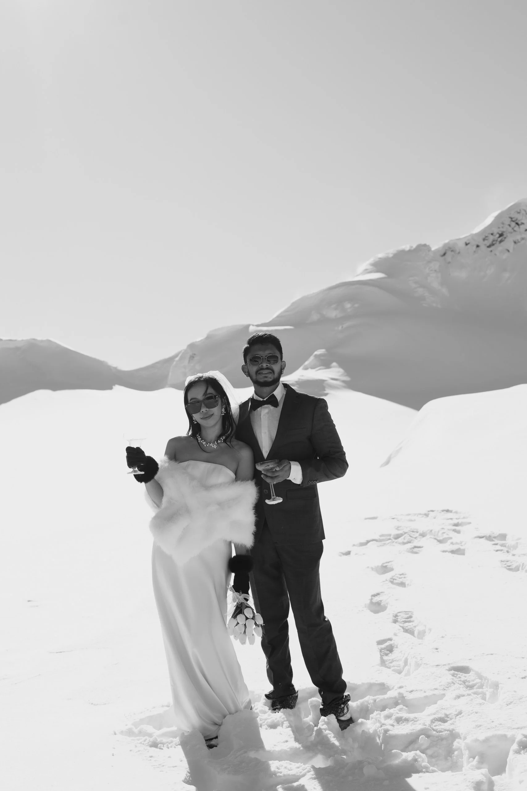 A couple dressed in formal attire standing in snow with snow-covered mountains in the background, holding glasses of champagne.