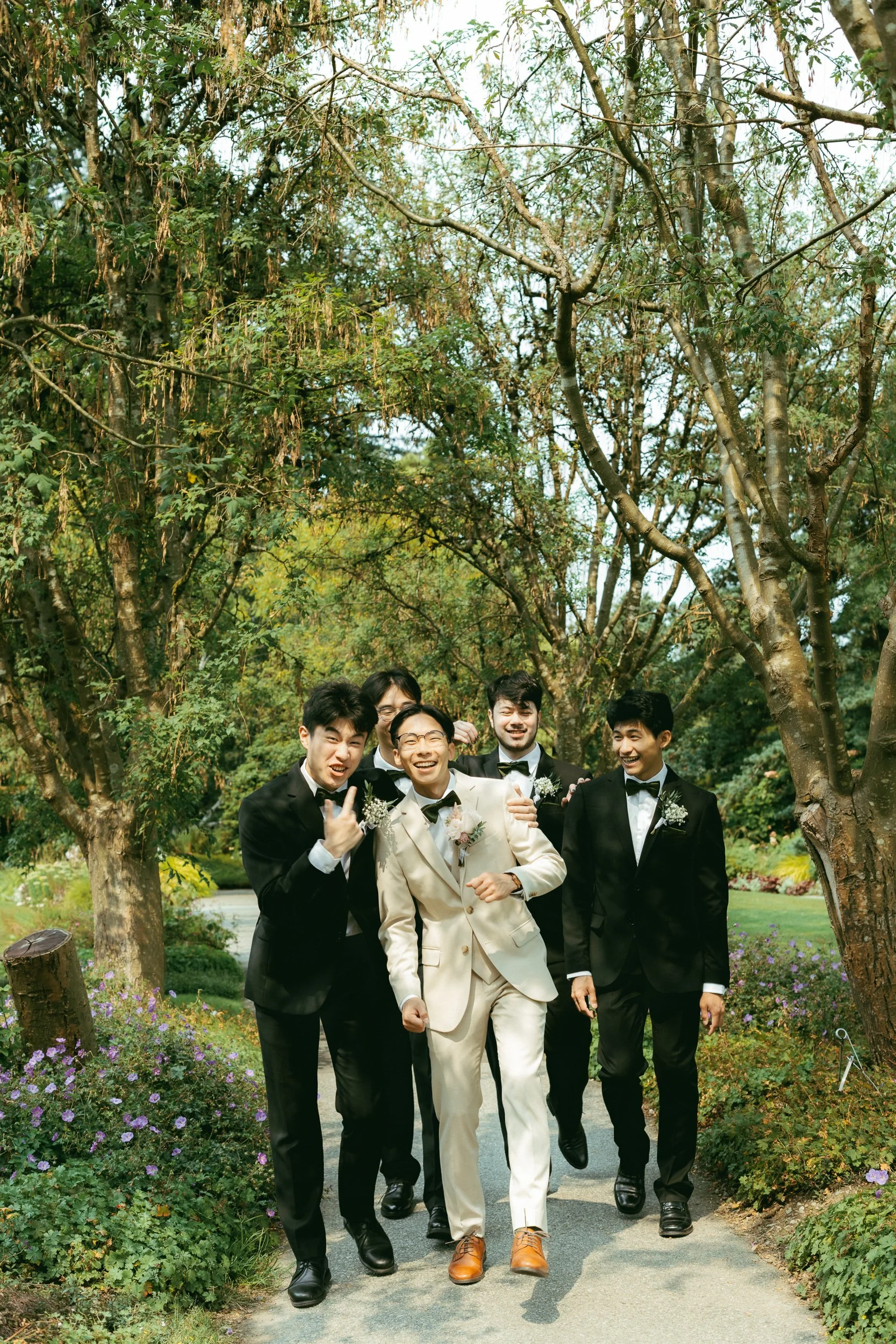 Group of young men dressed in tuxedos, with one in a cream-colored suit, walking and smiling along a garden path surrounded by trees and purple flowers.