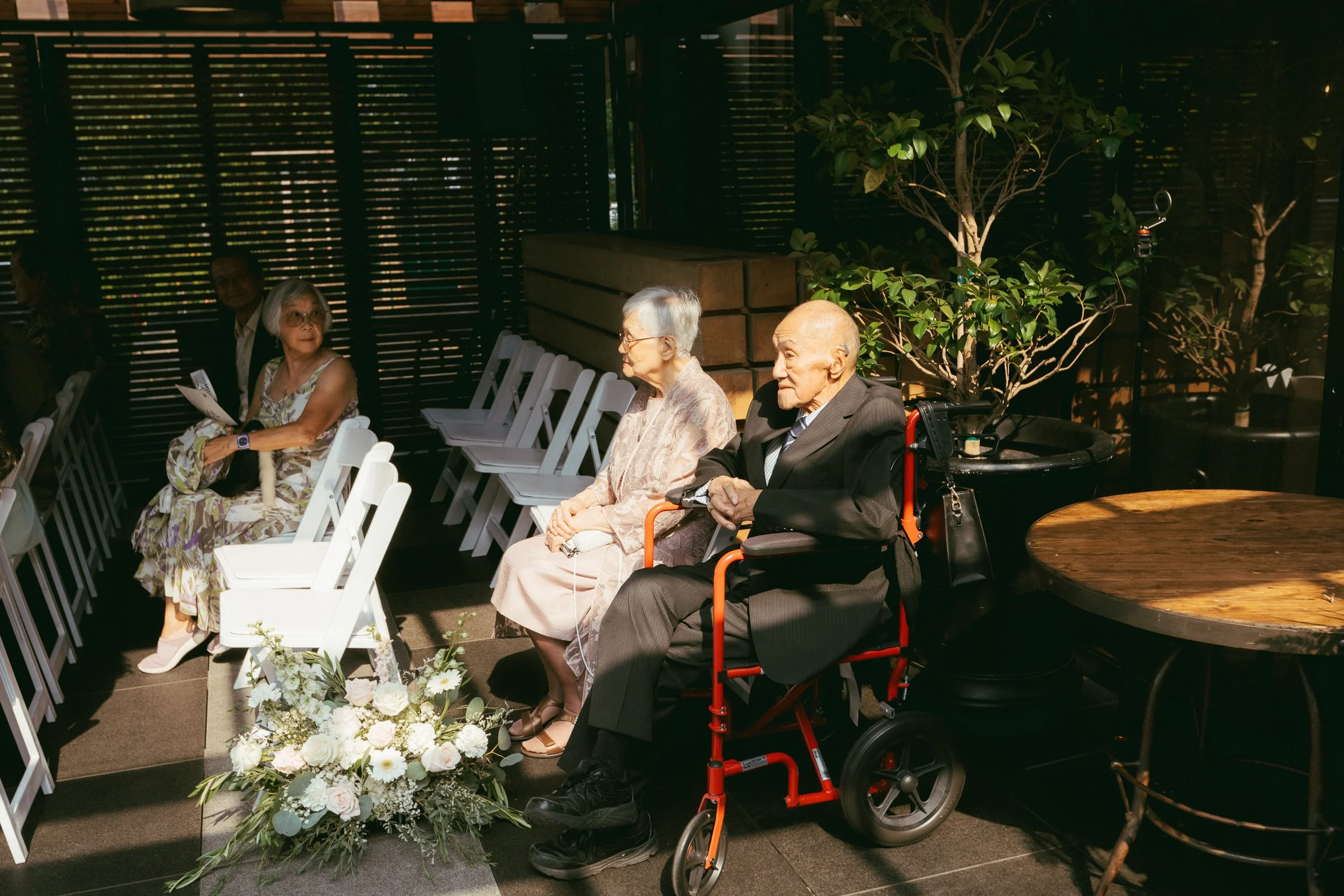 Elderly people sitting outdoors, some in chairs, one in a wheelchair, near a table with a floral arrangement, under a shaded structure with greenery.