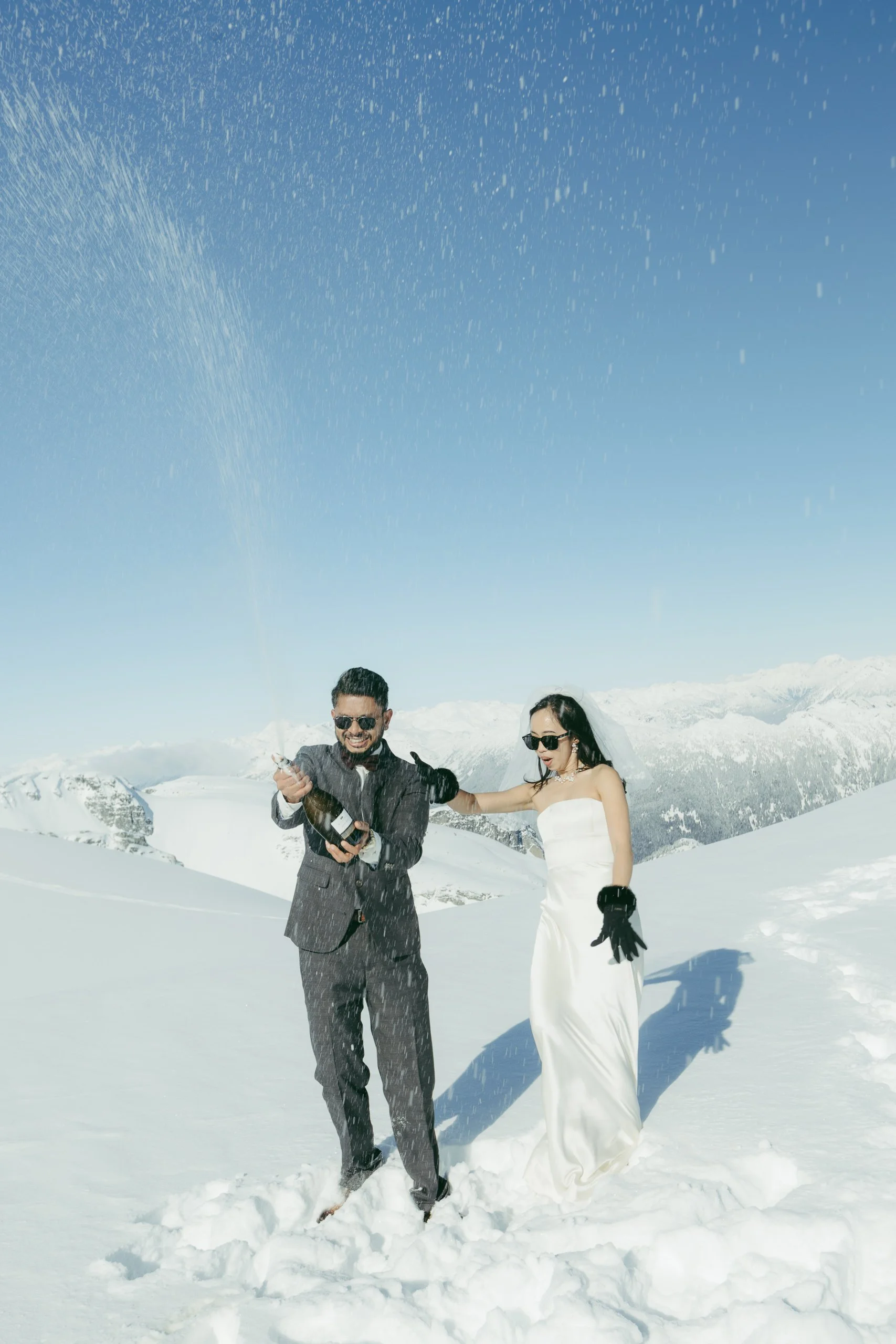 A bride and groom celebrating on a snowy mountain with champagne, the groom opening a bottle and the bride wearing a white wedding dress, sunglasses, and gloves, with snow-covered mountains in the background and blue sky above.