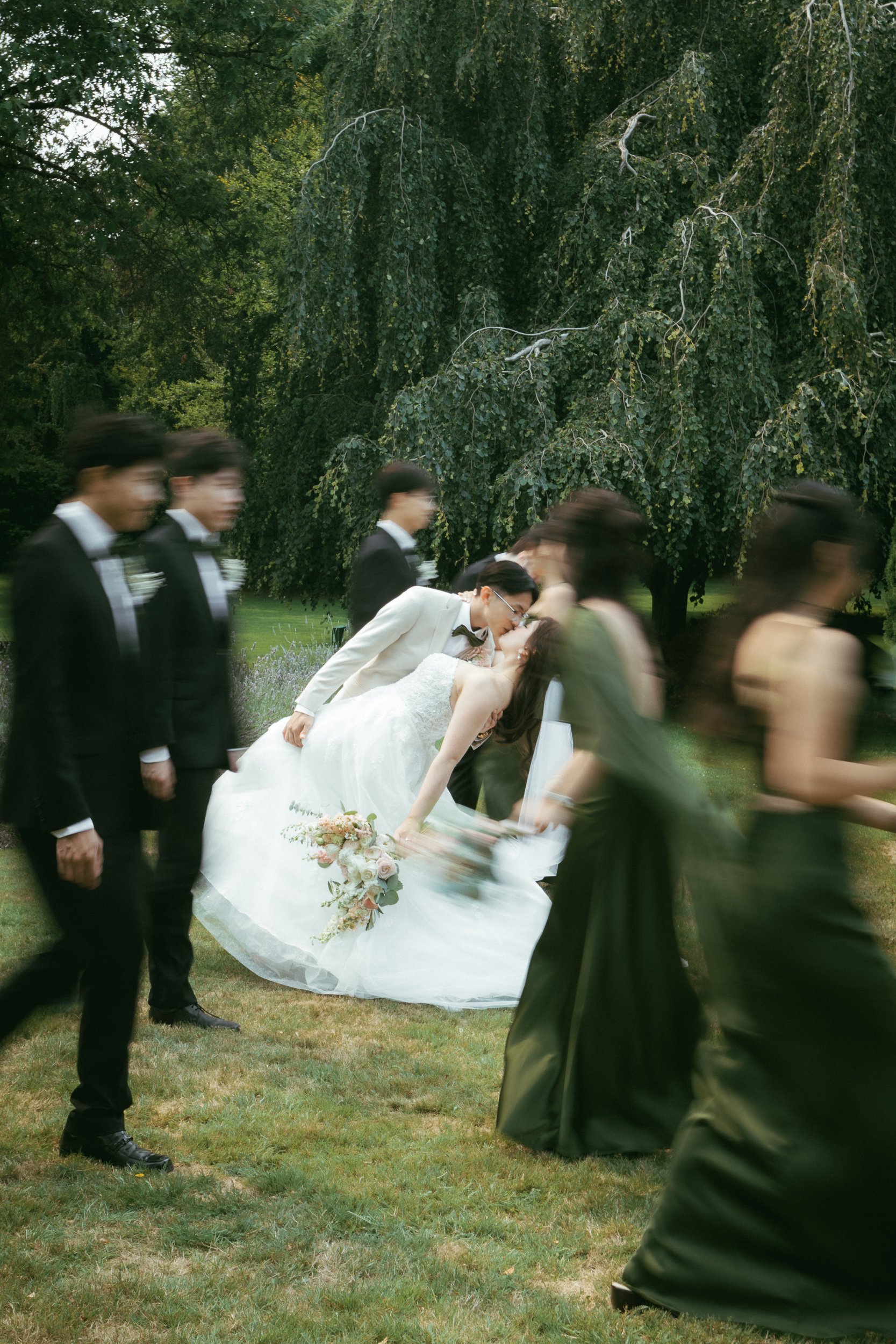 A bride and groom kissing, surrounded by wedding guests in formal attire, outdoors in a park with lush green grass and trees.