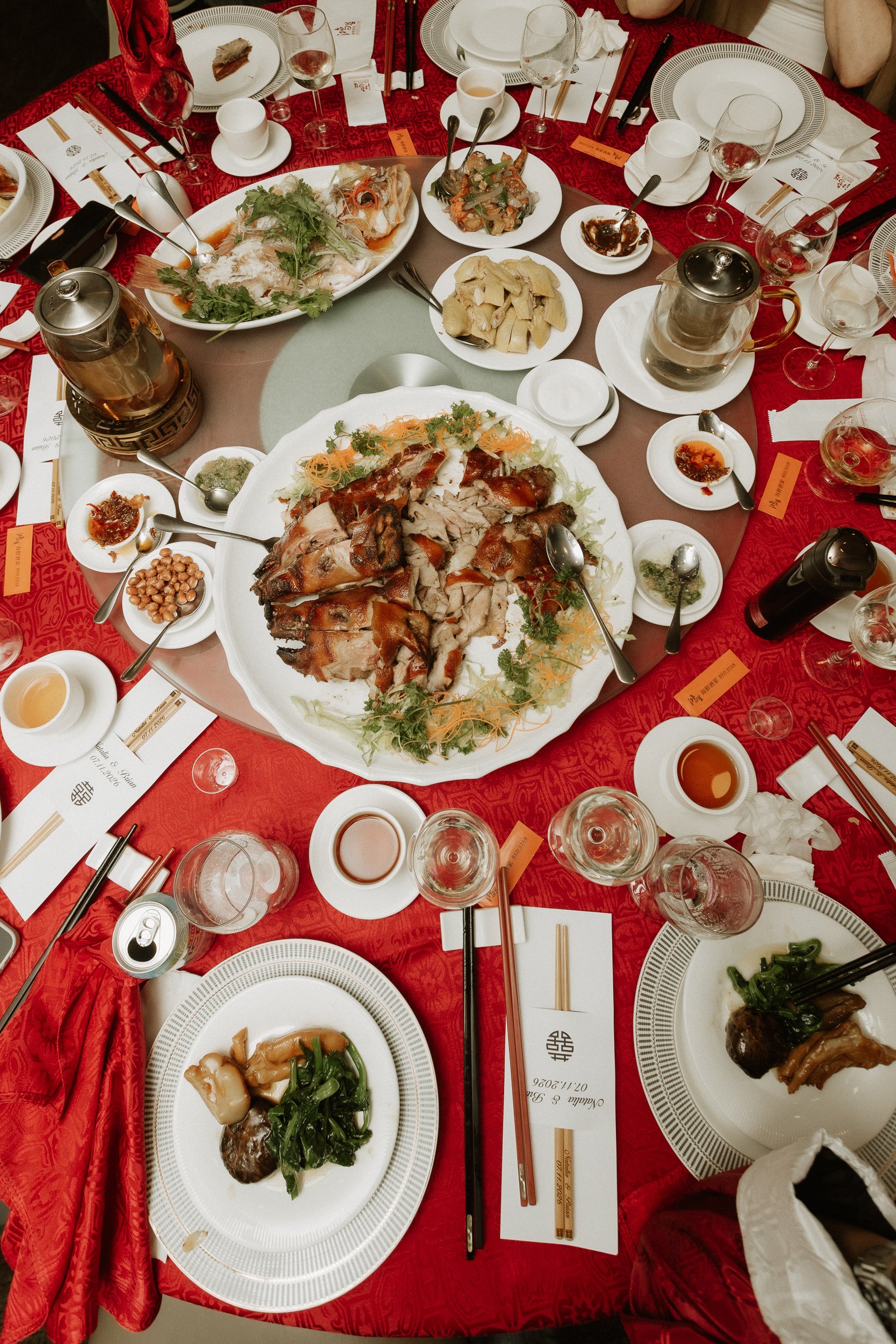 A round dining table set for a Chinese banquet, with various dishes including roasted chicken, fish, vegetables, and sauces, surrounded by empty plates, glasses, and chopsticks.