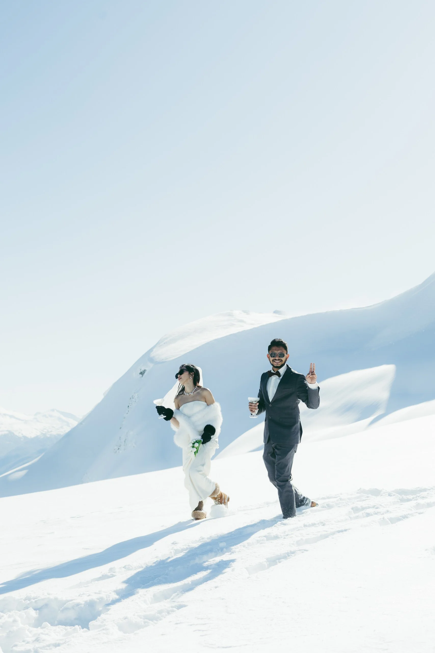 A man and woman in formal attire walking in the snow on a sunny day in a snowy mountain landscape. The man is wearing a tuxedo and sunglasses, holding a drink and making a peace sign. The woman is wearing a white fur shawl and dress, holding her phon