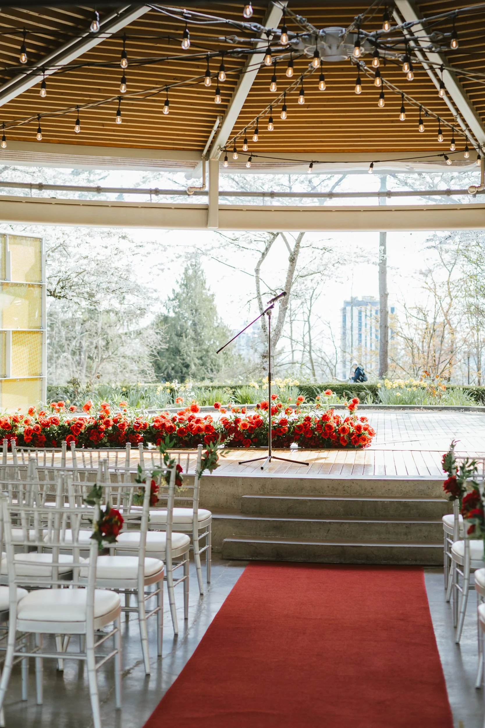 Indoor wedding or event venue with rows of white chairs adorned with red flower arrangements, a red carpet leading to a stage with a microphone, and string lights hanging from the ceiling, with a large window showing trees and a building outside.