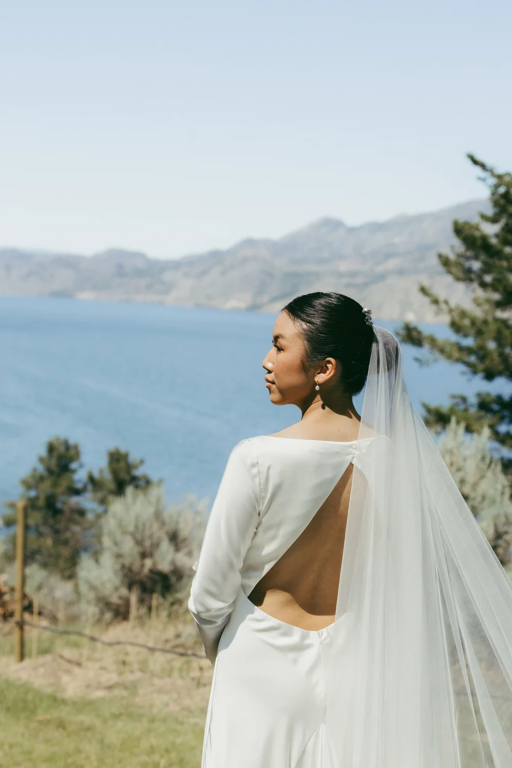 A bride in an open-back gown looking out over Okanagan Lake