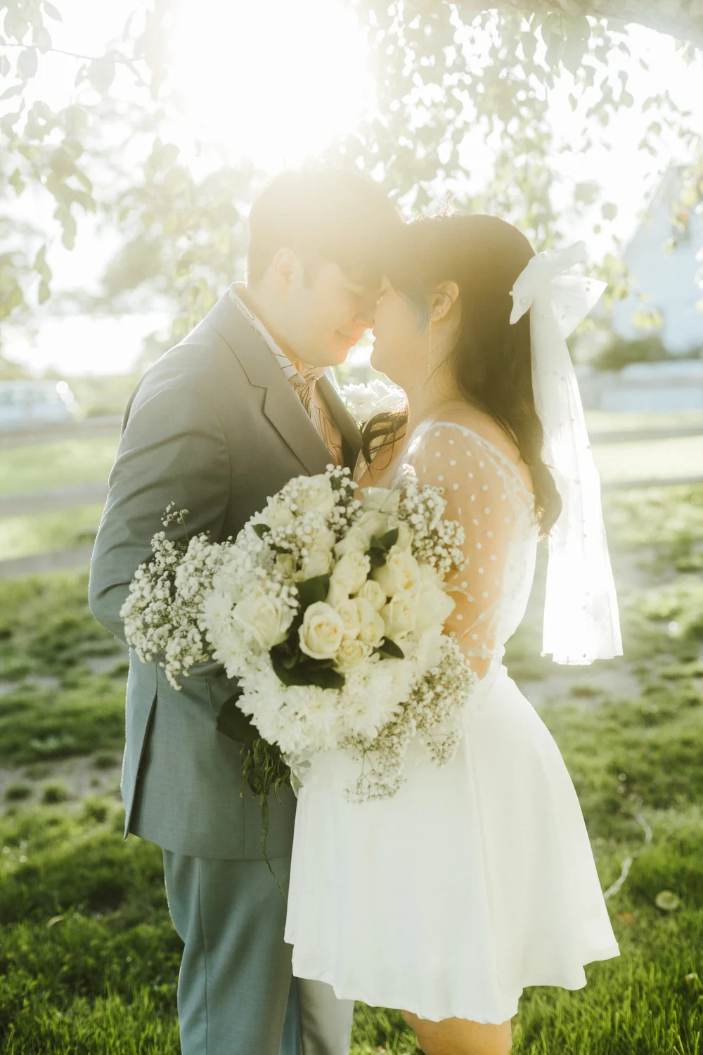 A couple kissing in soft backlight beneath blossoming trees, her bouquet of white roses between them