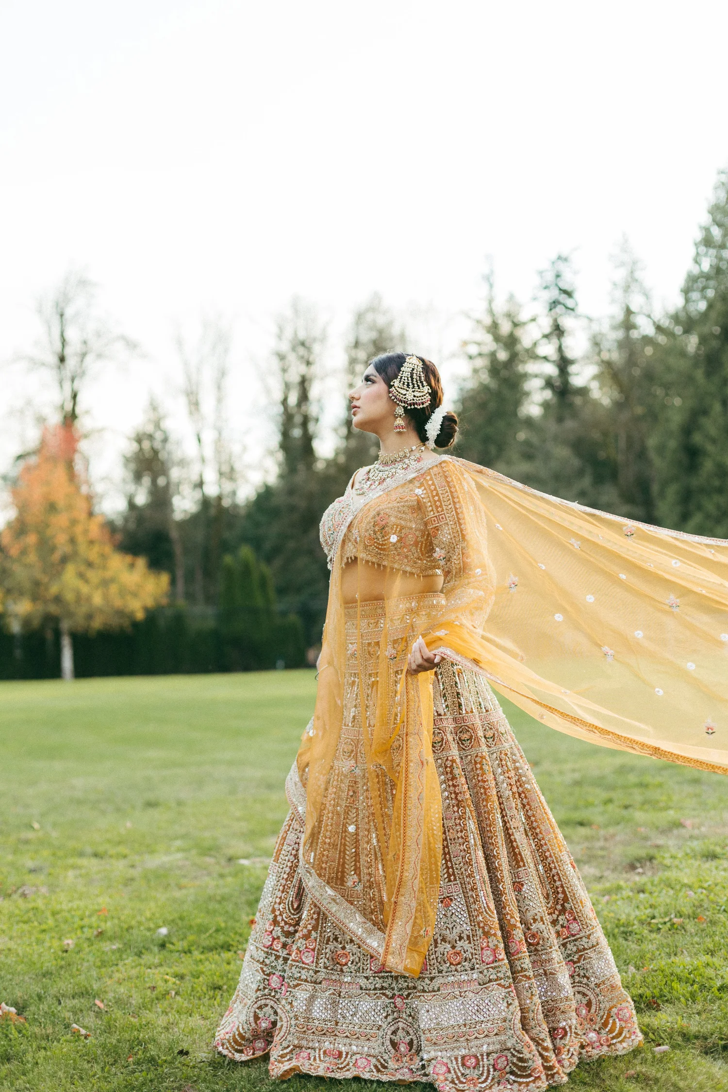 A South Asian bride in a golden lehenga, her dupatta lifting in the wind on a lawn