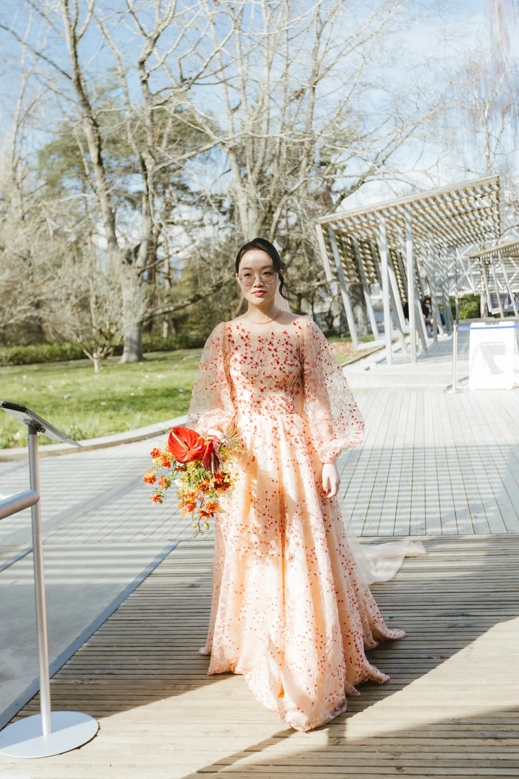 A woman wearing a peach-colored floral gown and glasses holding a bouquet of flowers on a wooden platform outdoors with bare trees and a modern architectural structure in the background.