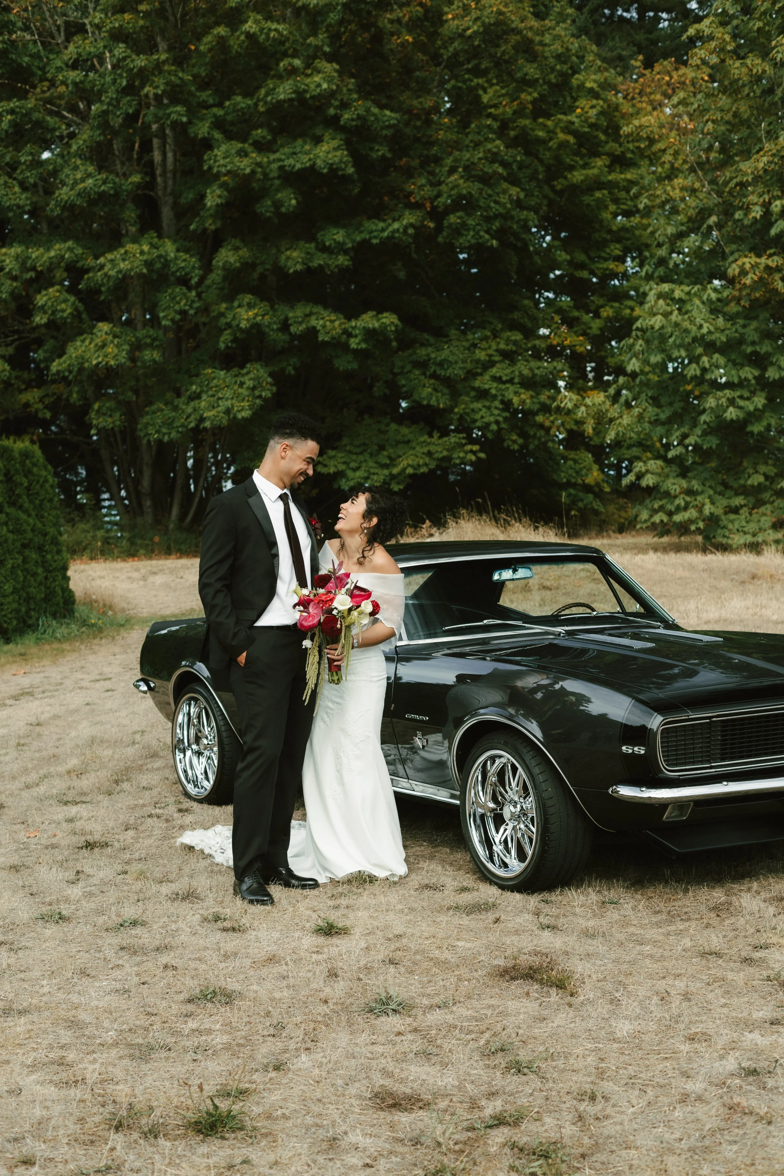 Smiling bride and groom with a colorful bouquet beside a classic black Chevrolet Camaro in a wooded field
