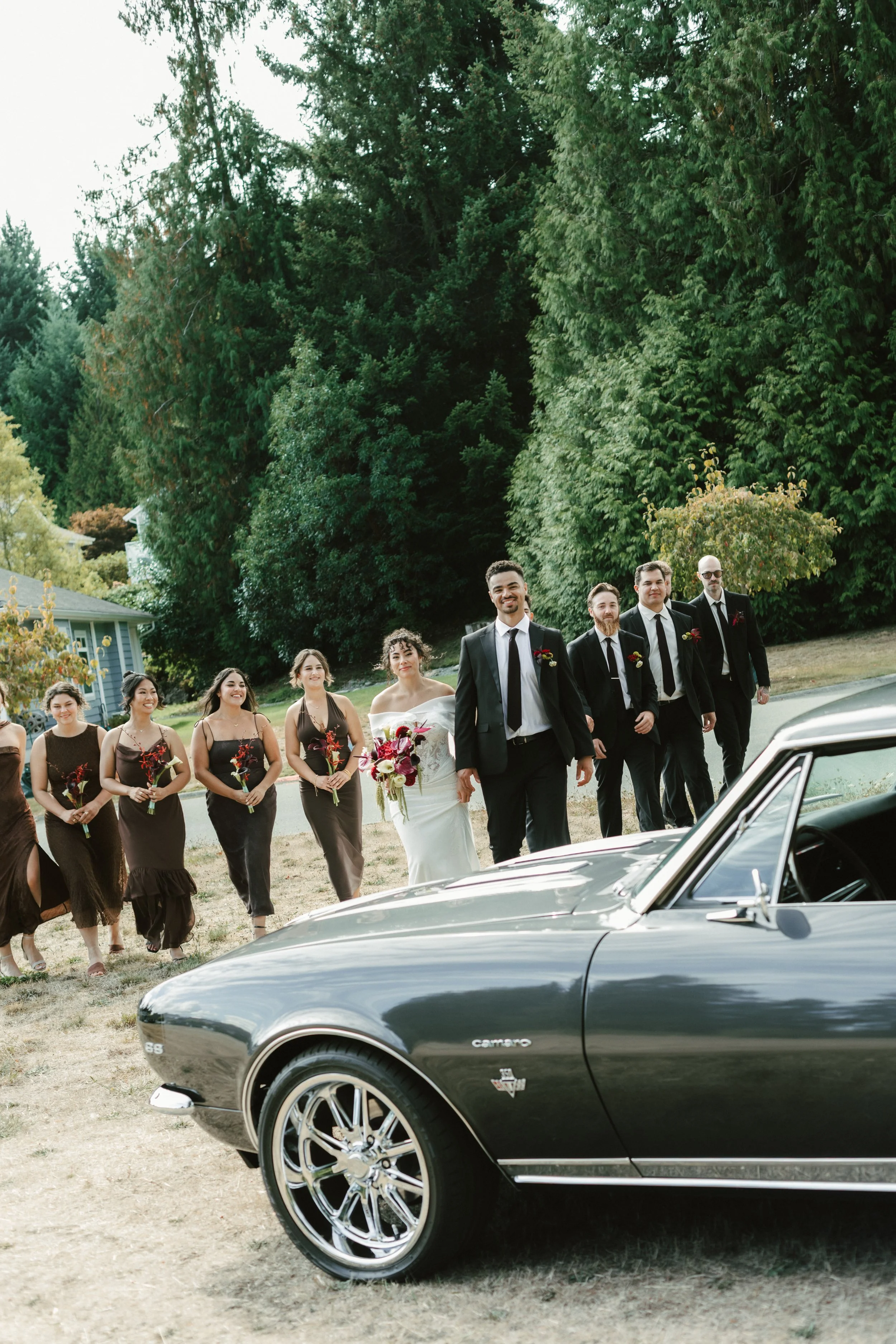 Wedding party gathered outdoors beside a classic black Chevrolet Camaro