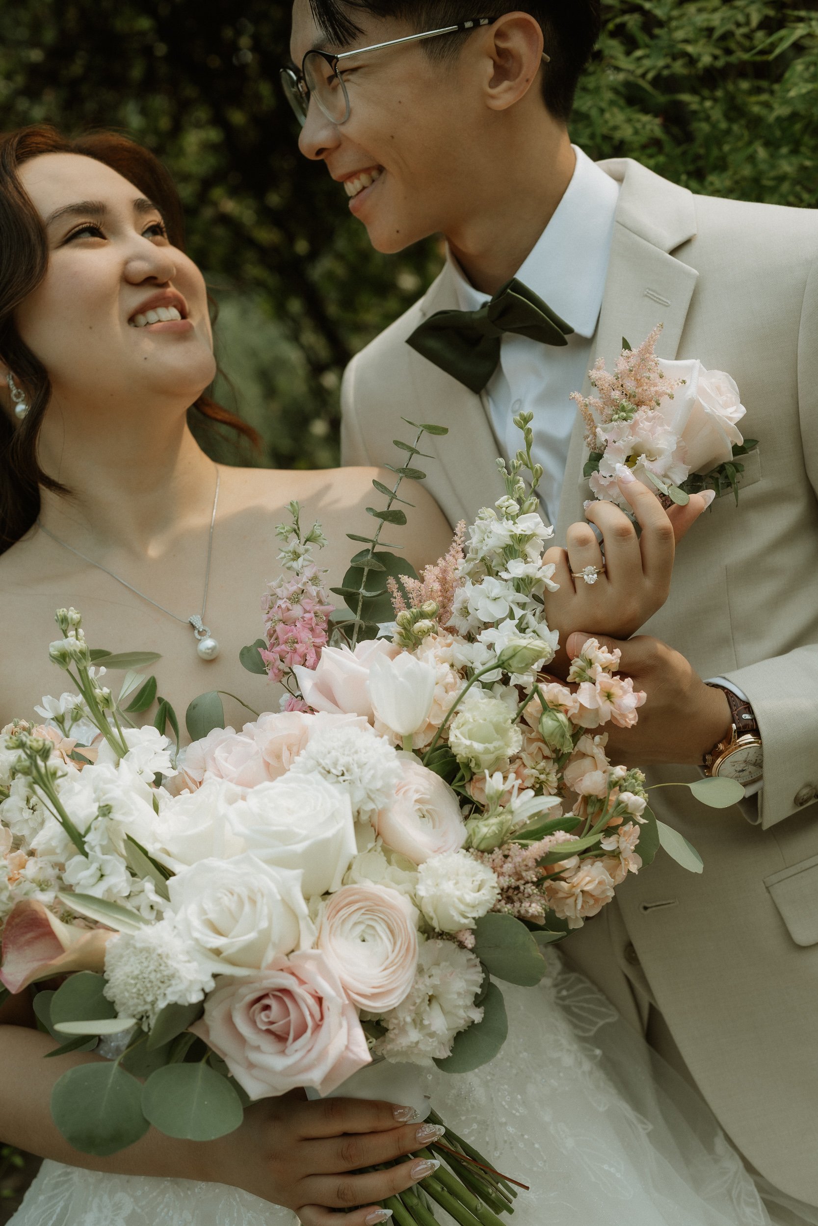 A bride and groom smiling at each other outdoors, holding a bouquet of pale pink and white flowers with greenery.